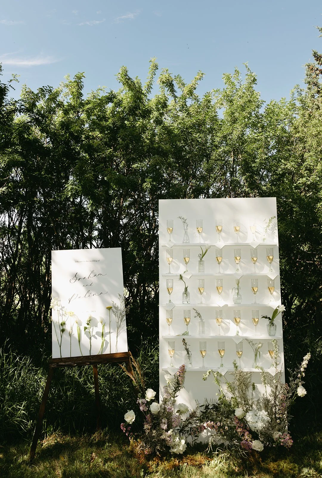 Wedding reception setup with a large white display board with flower arrangements and champagne glasses, surrounded by greenery and a clear blue sky. Forest wedding ceremony Calgary.
