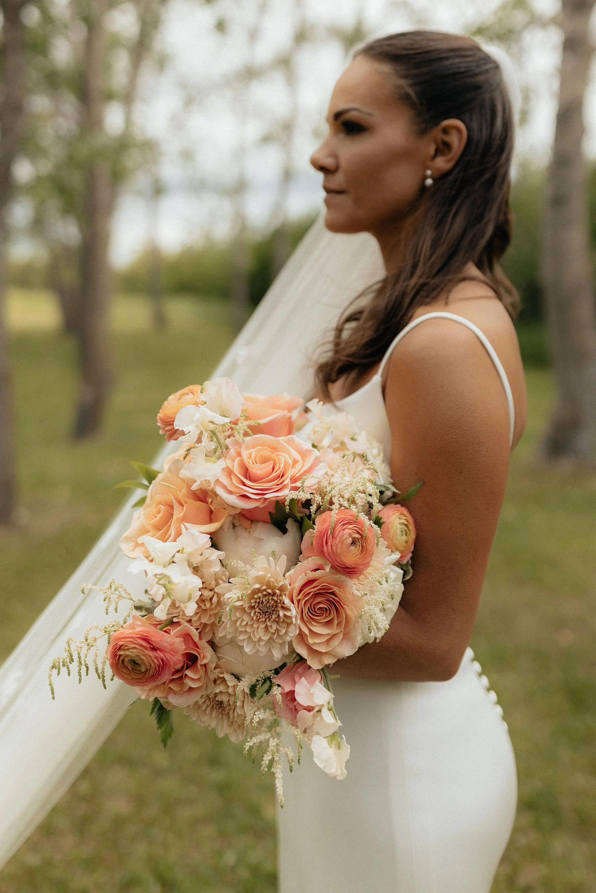 Bride holding a bouquet of peach and white flowers, standing outdoors with trees in the background at a Calgary Wedding Venue.