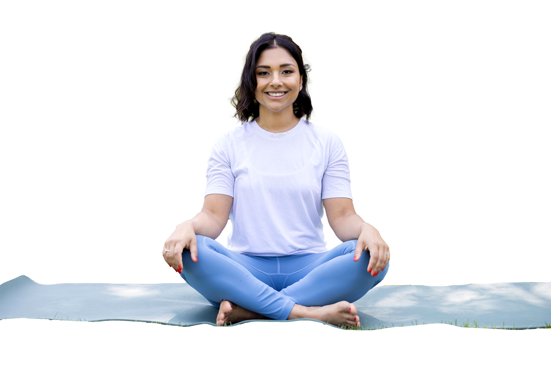 A woman sitting cross-legged on a yoga mat outdoors, smiling, dressed in a white t-shirt and blue leggings, practicing meditation.