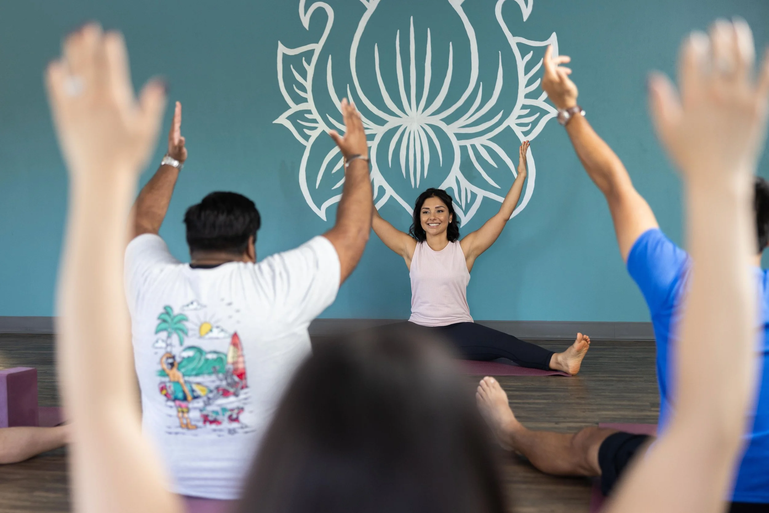 A woman leading a yoga class in a studio, sitting on a yoga mat with legs spread apart and arms raised, while students in the foreground mimic her pose with arms raised as well. The studio has a large decorative wall art of a lotus flower behind the instructor.