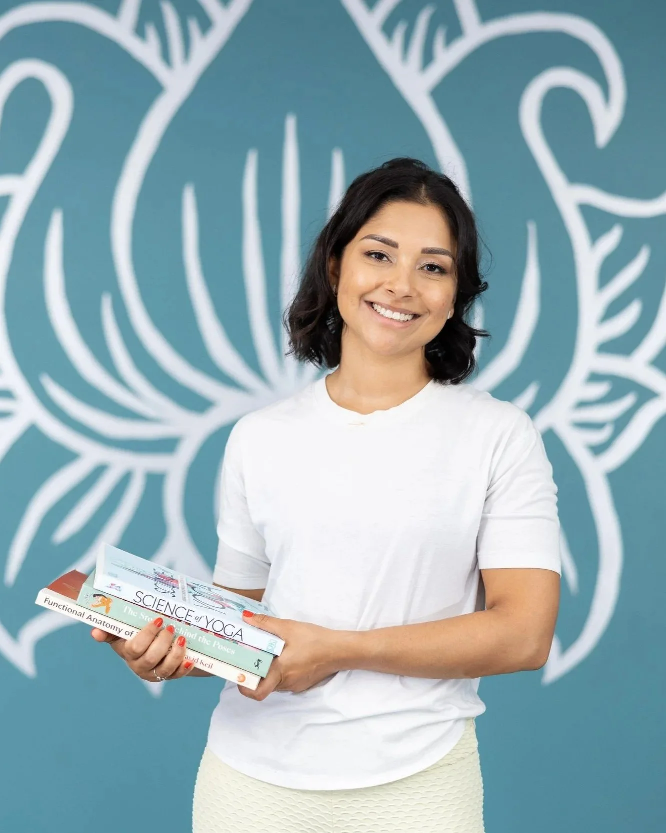 A woman with dark hair and a bright smile holding several books, standing in front of a teal wall with a large white lotus flower design.