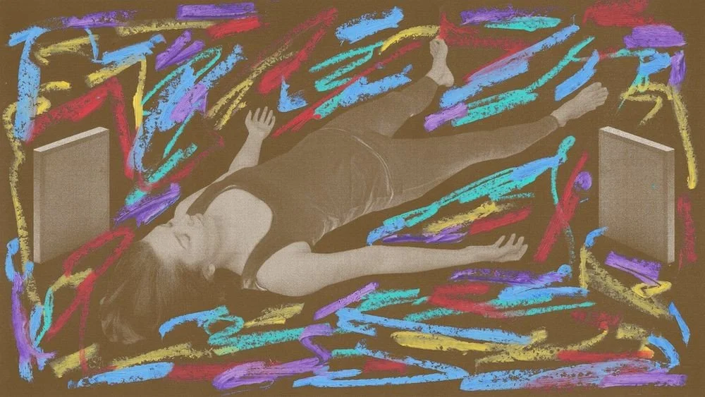 A woman lying on the floor surrounded by colorful chalk scribbles with two speakers nearby.