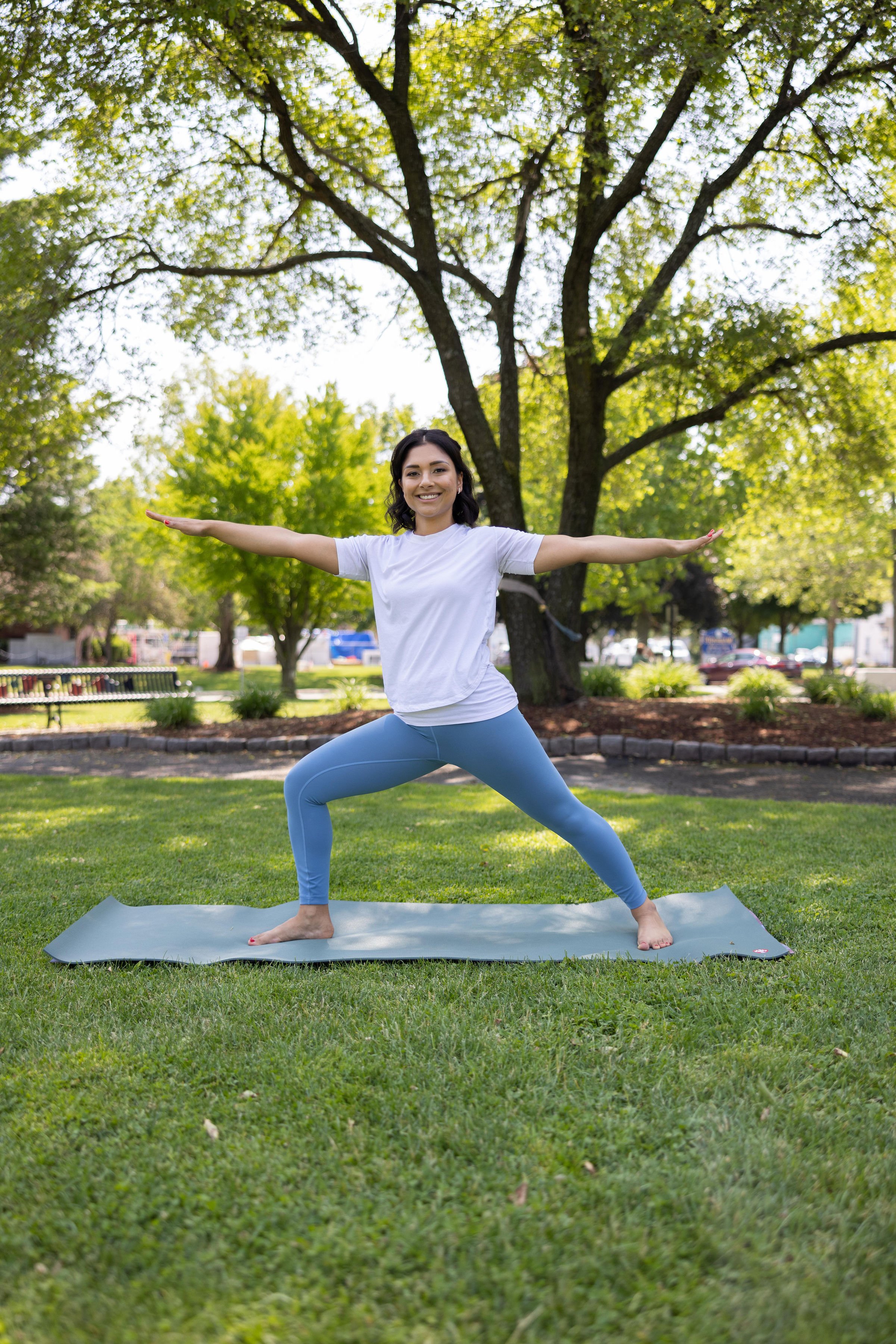 Woman practicing yoga outdoors on a gray mat in a park, standing in a warrior pose with arms extended to the sides, smiling, with trees and greenery in the background.