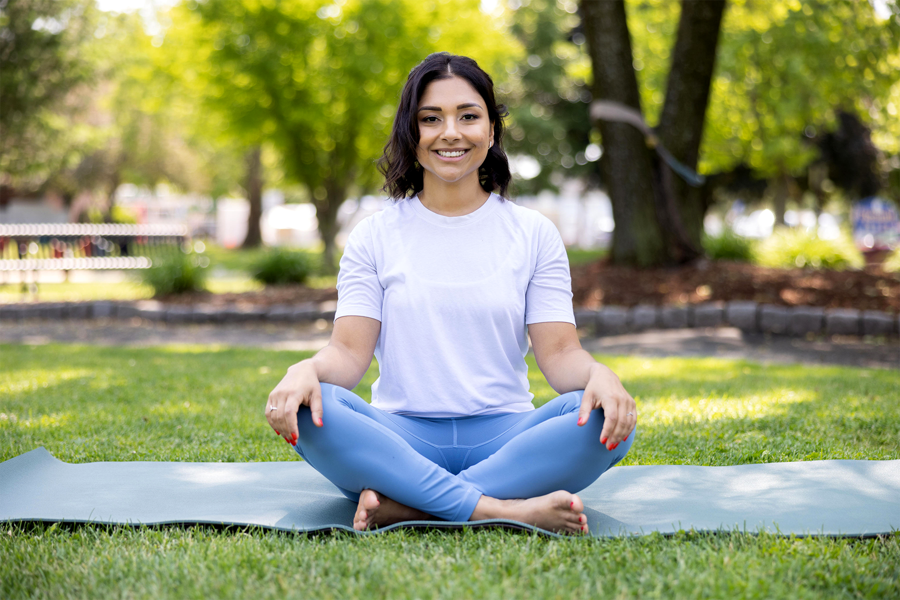 A woman sitting cross-legged on a yoga mat outdoors, smiling, in a park with green grass and trees in the background.