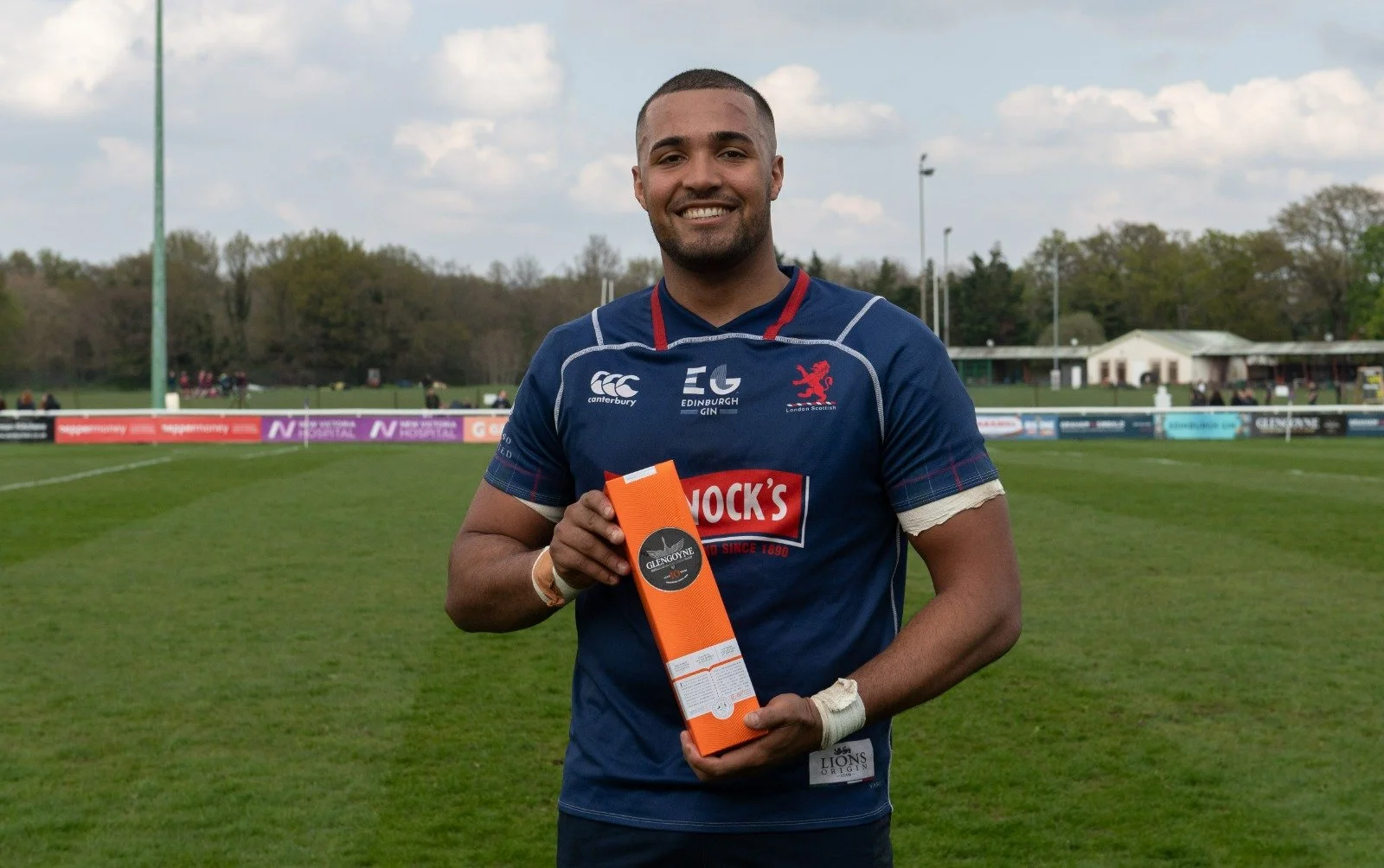 Smiling rugby player in blue jersey holding an orange Glengoyne whiskey box on a grassy field.