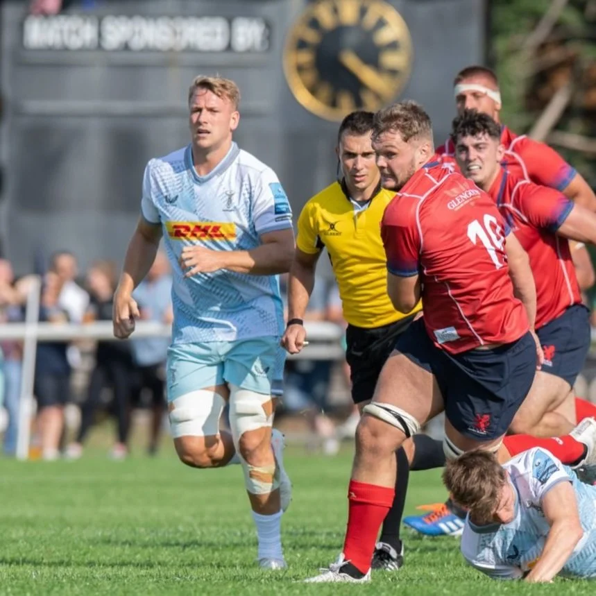 Rugby players in action during a match, with one player in a blue jersey and another in a red jersey. A referee in a yellow shirt is visible in the background.