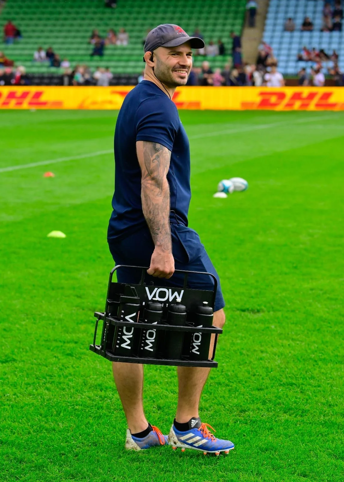 A man in athletic clothing standing on a rugby field, holding a water bottle carrier with several water bottles, and smiling.