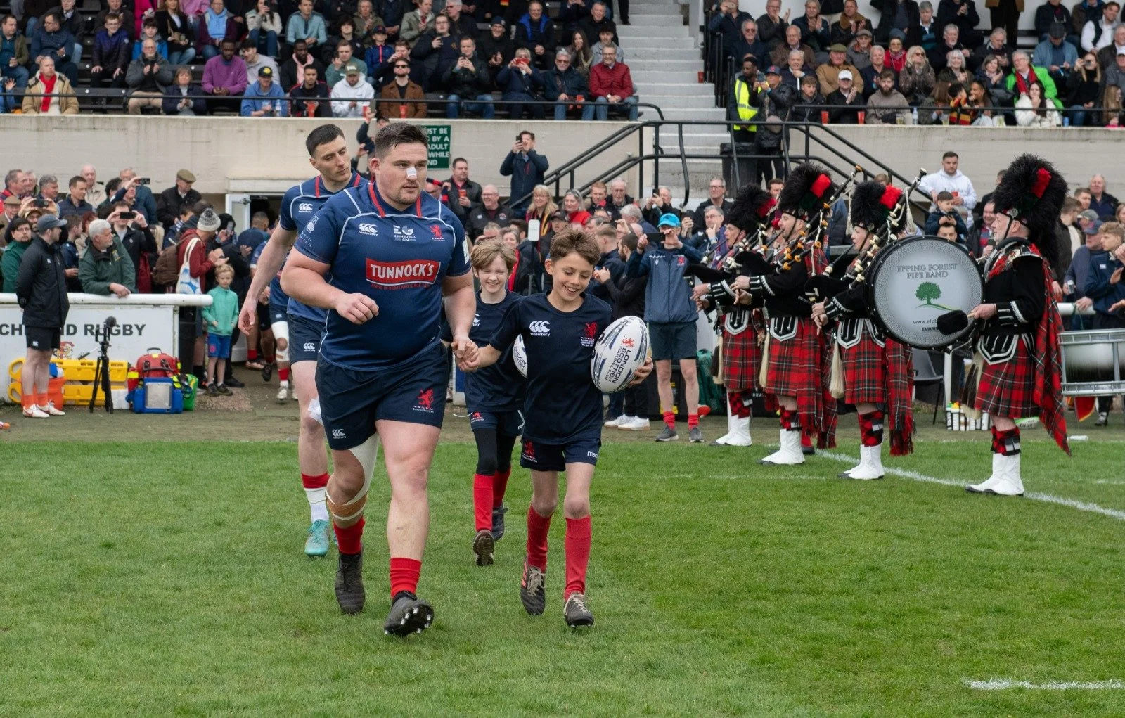 Rugby players walking with a child carrying a rugby ball on a field, surrounded by a bagpipe band in Scottish attire, with a crowd watching.