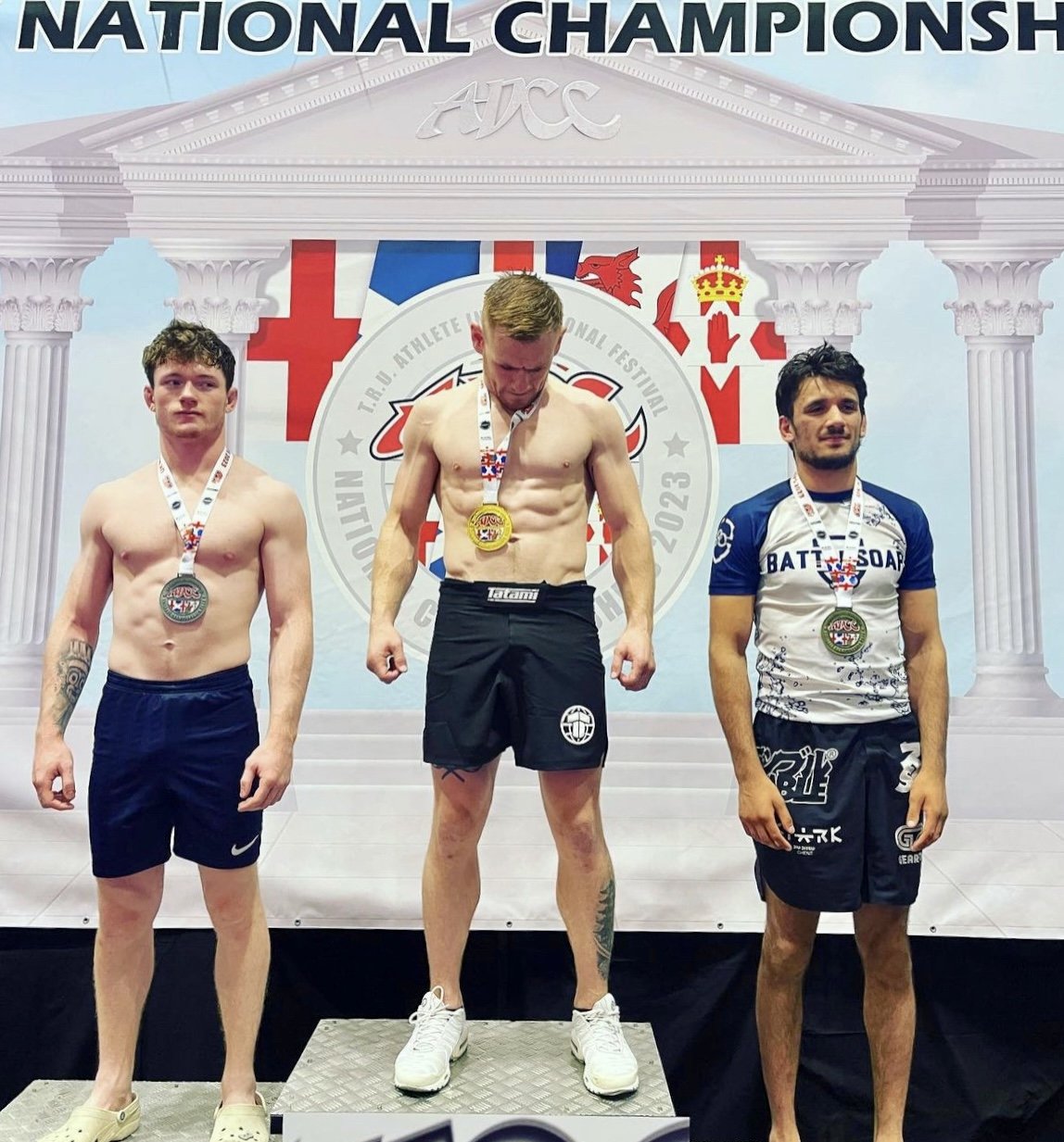Three athletes on a podium at the ADCC National Championship. The center athlete is wearing a gold medal, the left athlete has a silver medal, and the right athlete has a bronze medal. "ADCC" and flags are visible in the background, and the athletes are in athletic gear.