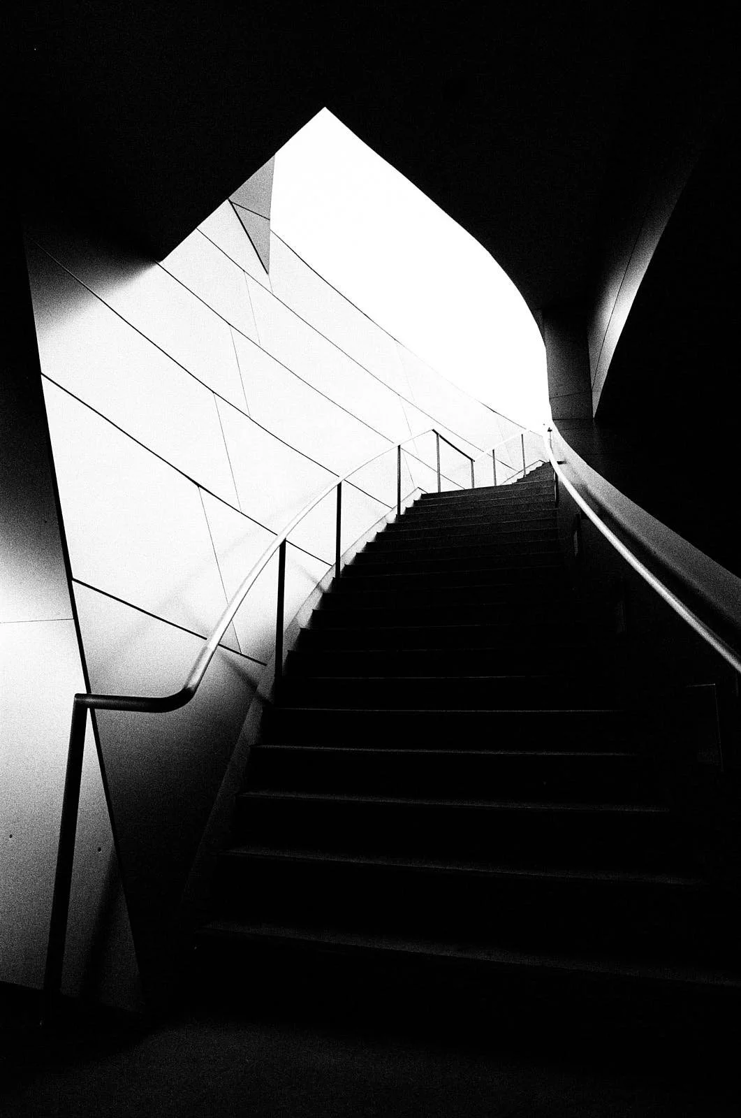 Black and white photo of a staircase with metal handrails, leading upward between curving walls and ceiling.