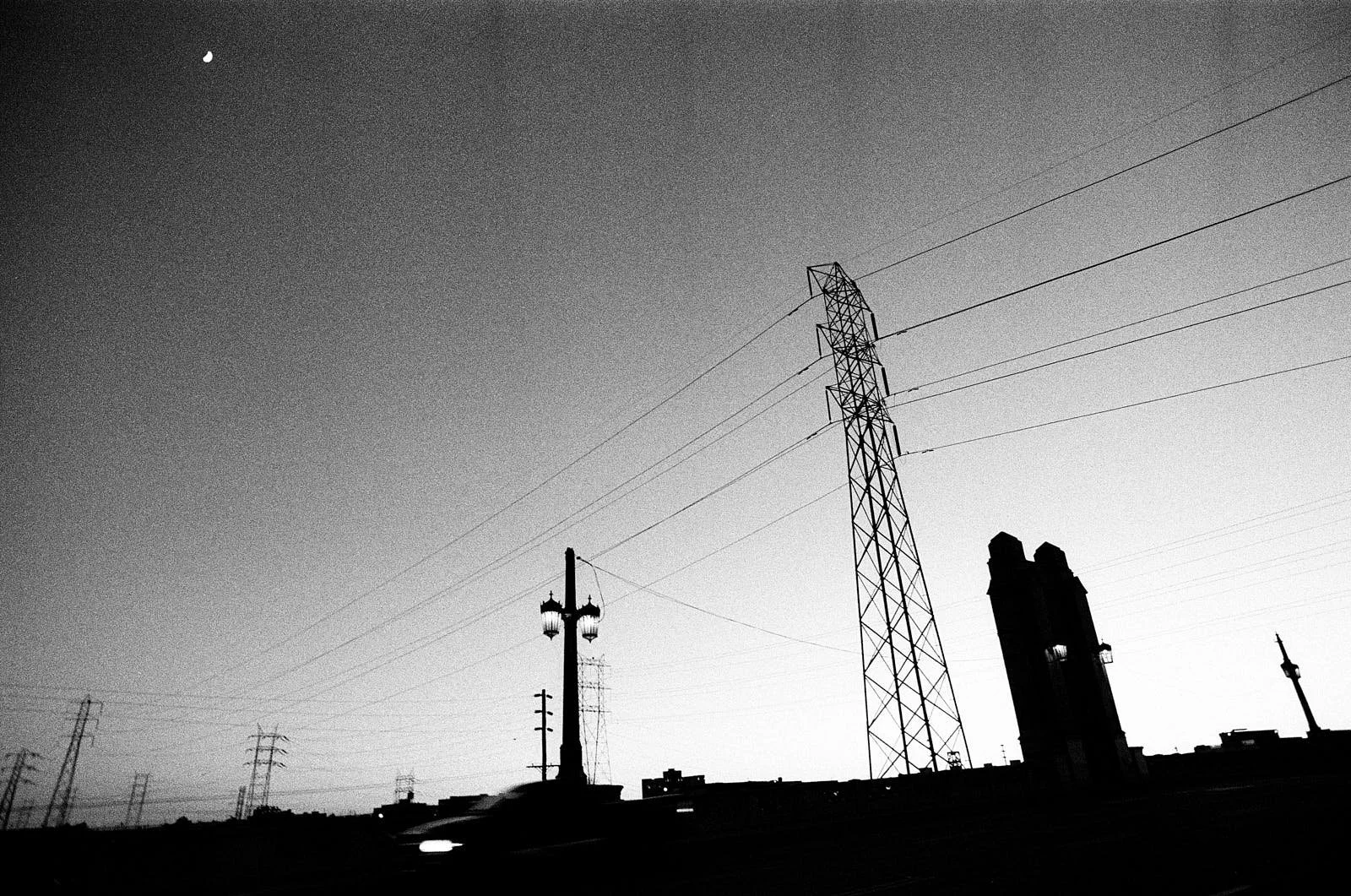 Black and white photograph of a cityscape at sunset with power lines and communication towers silhouetted against the sky.