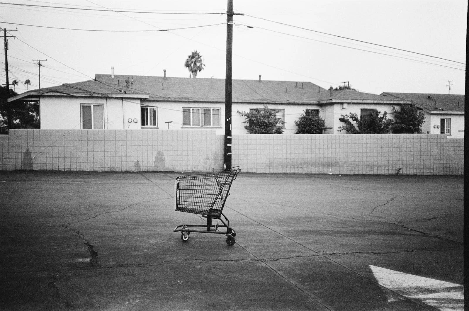 Empty shopping cart in an open parking lot, with a background of residential houses separated by a fence and visible power lines overhead.