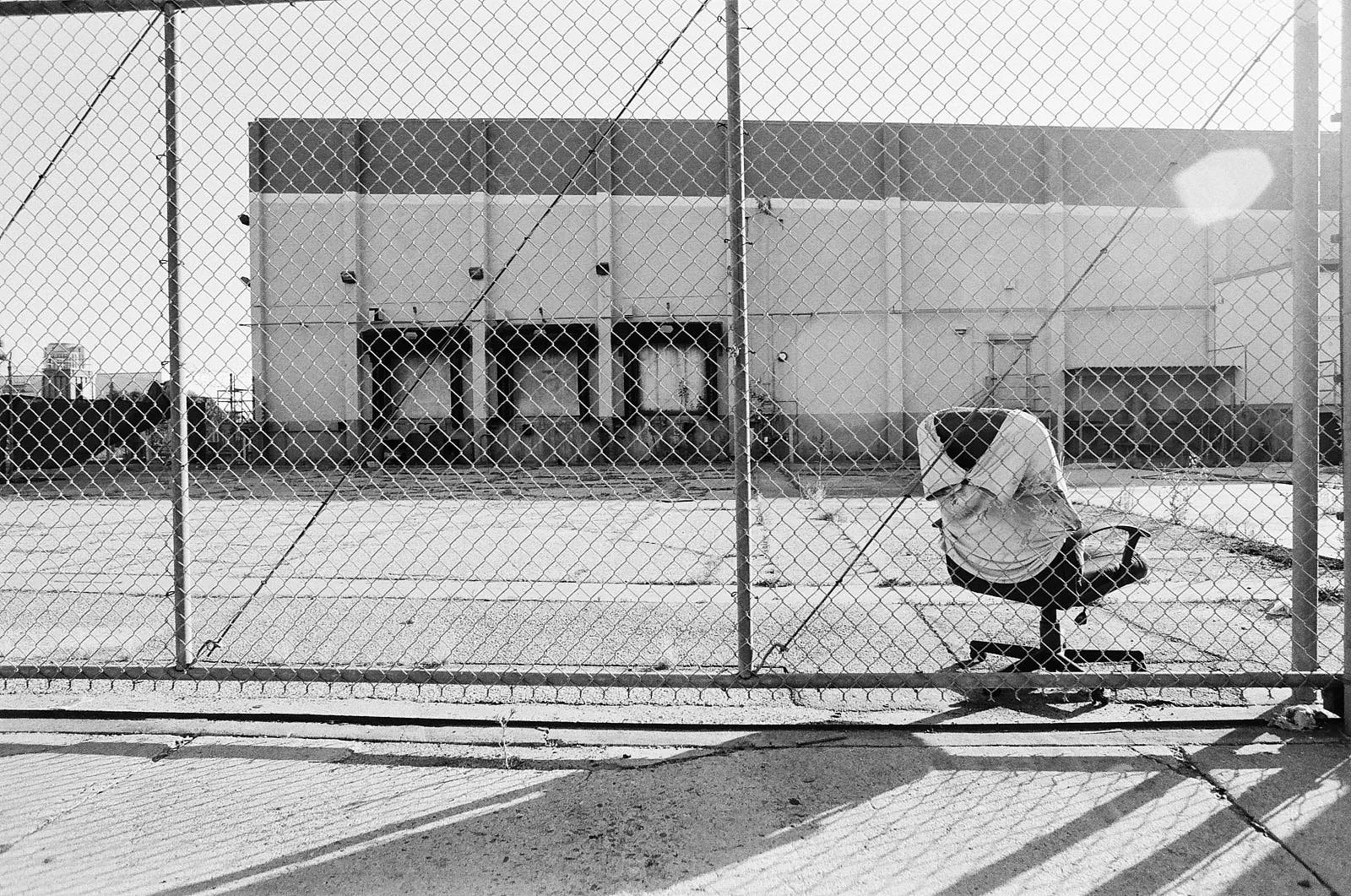 A person crouching on a rolling office chair behind a chain-link fence with a large industrial building in the background on a sunny day.