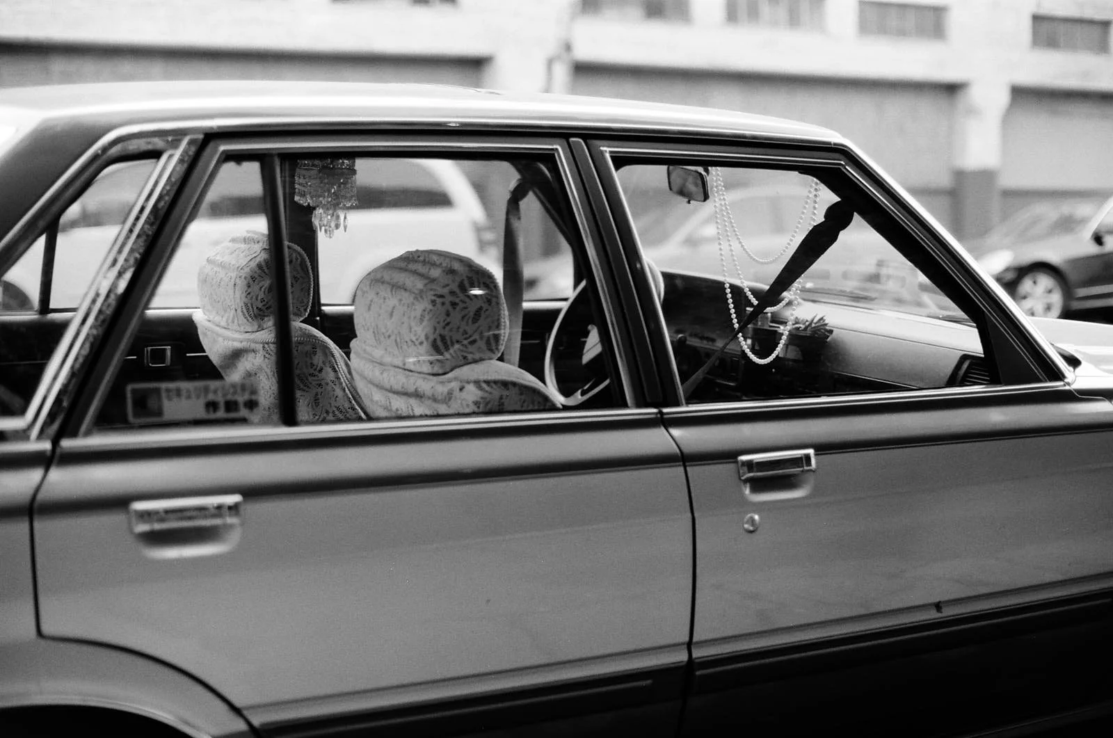 Black and white photo of an old car with lace seat covers, decorative necklaces hanging inside, parked in a lot with a building in the background.