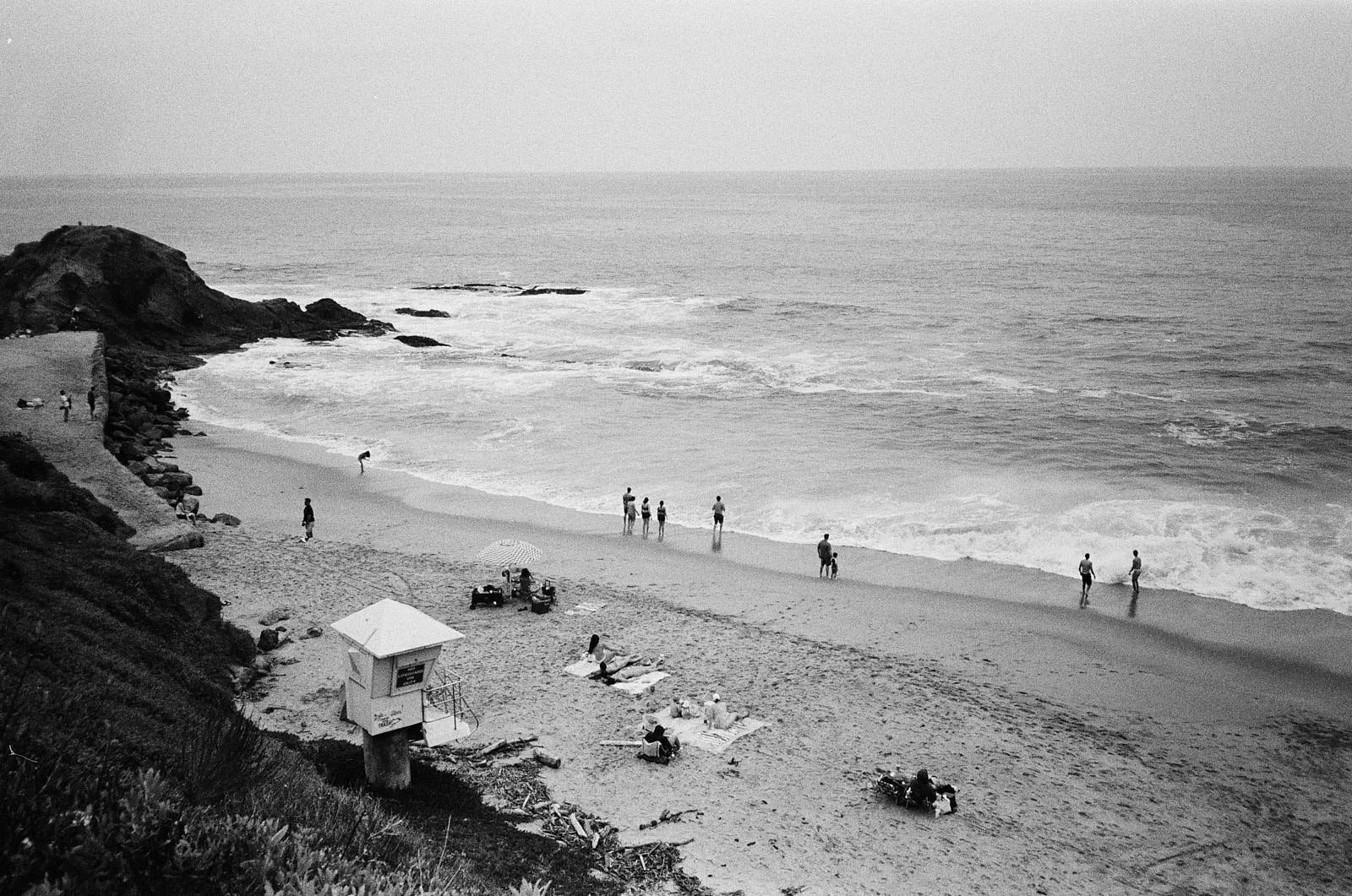 A black and white photo of a beach with several people walking along the shoreline, sitting on towels, and near a lifeguard station, with a rocky cliff on the left side and the ocean in the background.