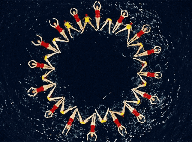 A group of synchronized swimmers forming a circle pattern in the water, wearing red and yellow swimsuits.