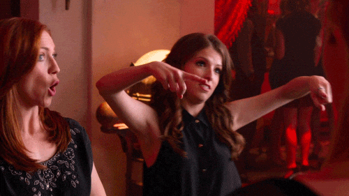 Two women sitting at a table in a dimly lit room, one with red hair and the other with brown hair, making a hand gesture as if pointing or demonstrating something, with a background of people and red lighting.