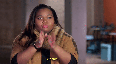 A young woman with long, dark hair wearing a brown and black jacket makes a gesture with her hands in front of her face, with the caption 'Boom!' at the bottom.