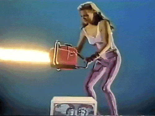 A woman with a long hairstyle wearing a tank top and striped pants holding a chainsaw that is emitting fire.