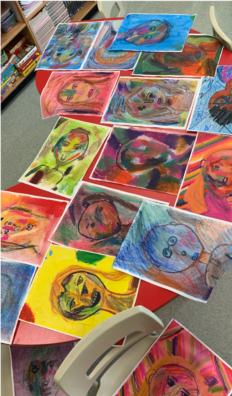 Colorful children's art portraits displayed on a red table, with some sketches on watercolor paper, featuring abstract and expressive faces created with various bright colors.