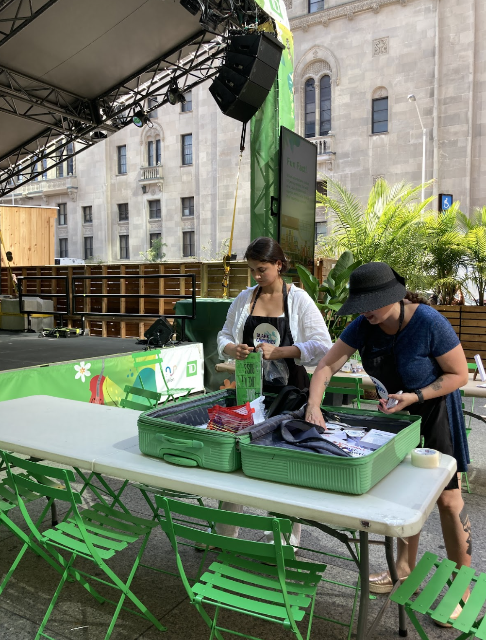 Two women setting up a table with green baskets at an outdoor event. One woman is wearing a black hat and a dark dress, while the other is in a white shirt. The event appears to be related to Earth Day, as indicated by the decorations and signage.