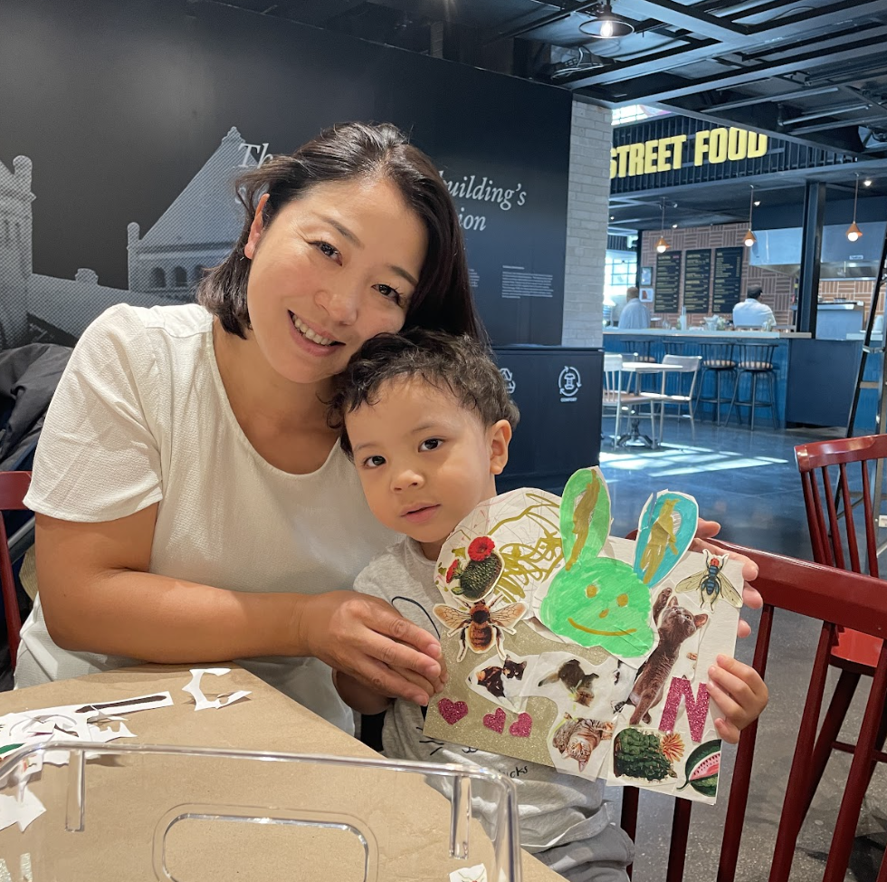 A woman and young boy sitting at a table in a restaurant or food court, with the woman smiling and the boy holding a colorful craft project decorated with drawings and stickers of animals, hearts, and nature themes.