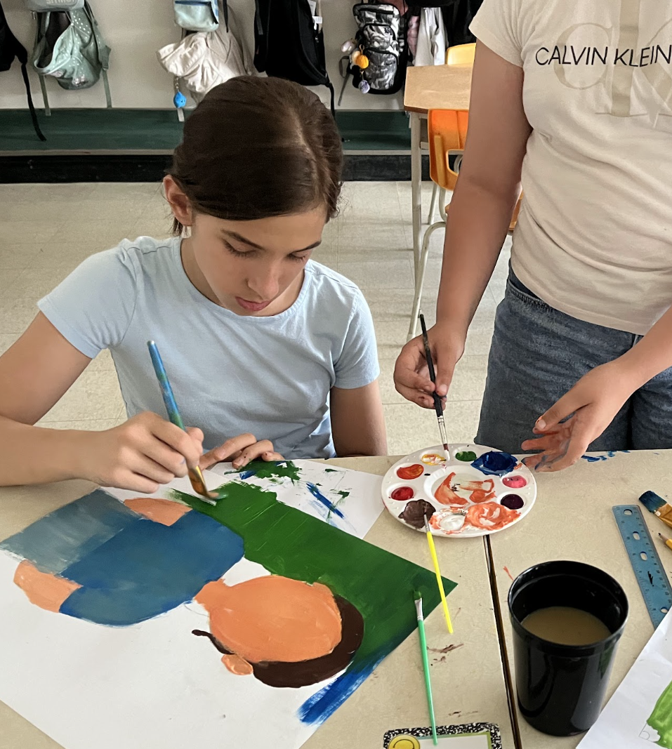 Young student with paintbrush painting a green background on a white board with a paint palette and wash cup beside her