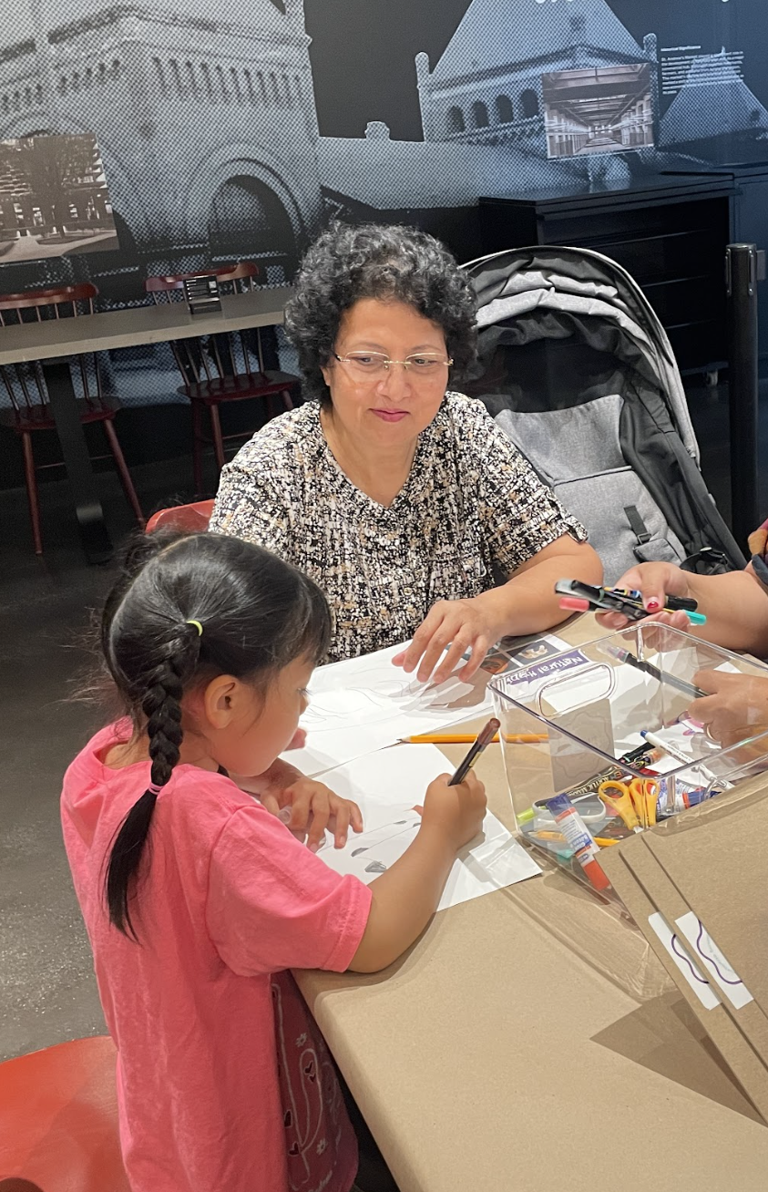 A woman and a young girl seated at a table, engaging in arts and crafts activities with markers, pencils, and glue, in an indoor setting with a gray wall and picture mural in the background.
