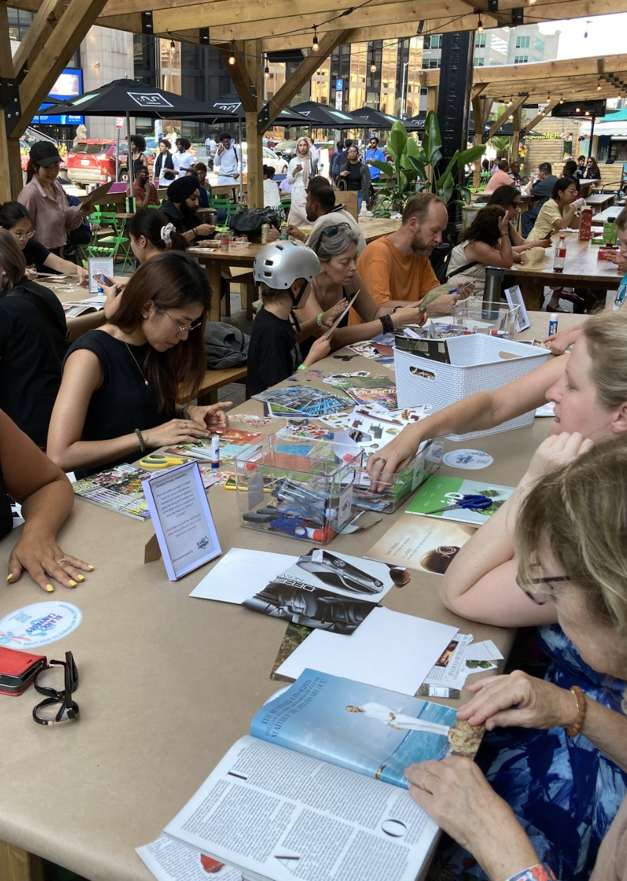 People sitting at a long table during a crafting event, with urban buildings and outdoor seating in the background.