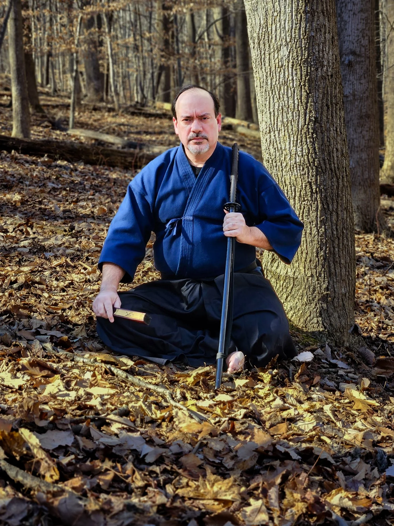 A man dressed in a blue martial arts uniform kneeling on a bed of fallen leaves in a wooded area, holding a sword in his left hand.