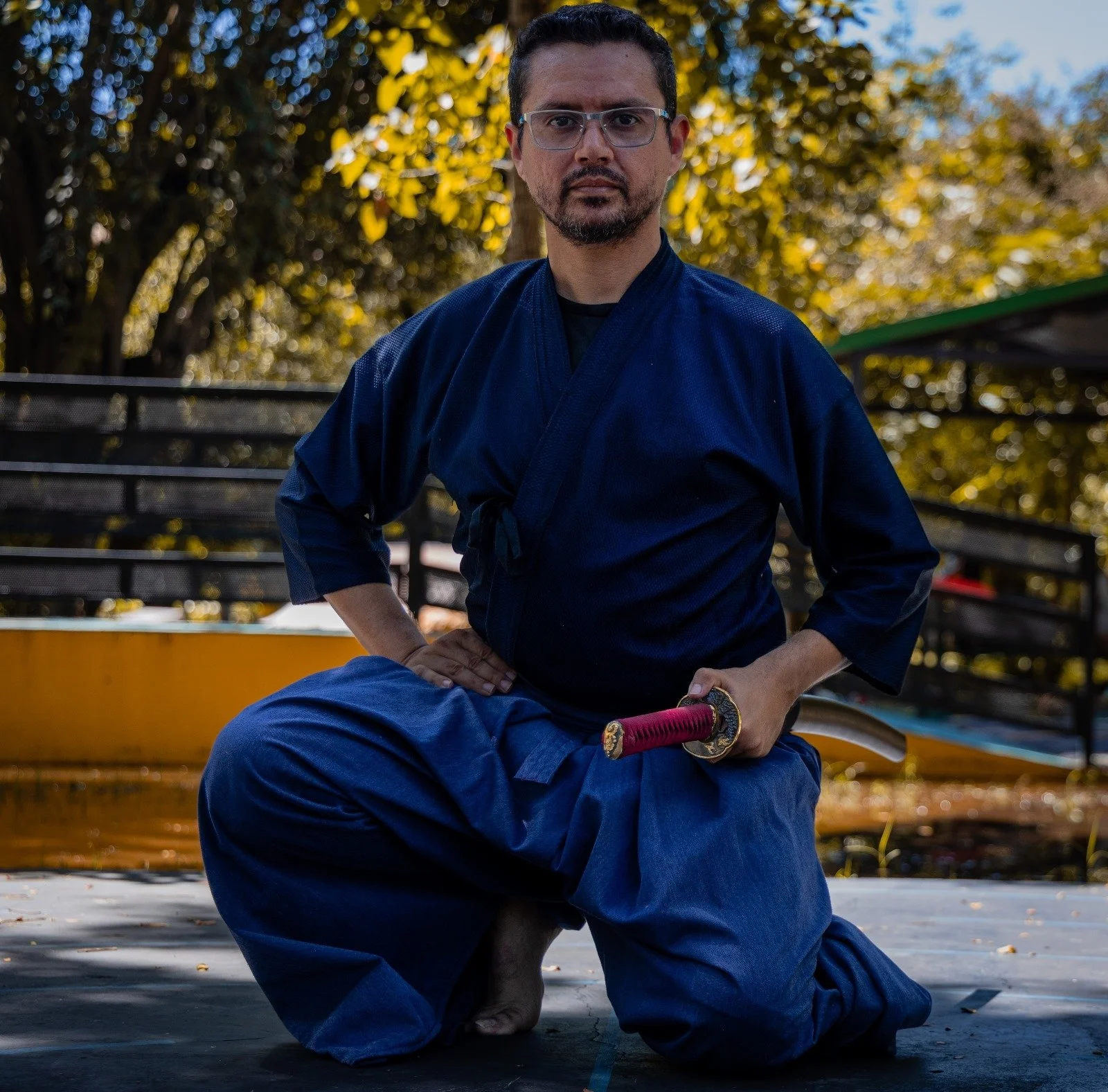 A man dressed in a traditional martial arts uniform, kneeling outdoors on a mat, holding a katana sword with a red handle, with trees and a park setting in the background.