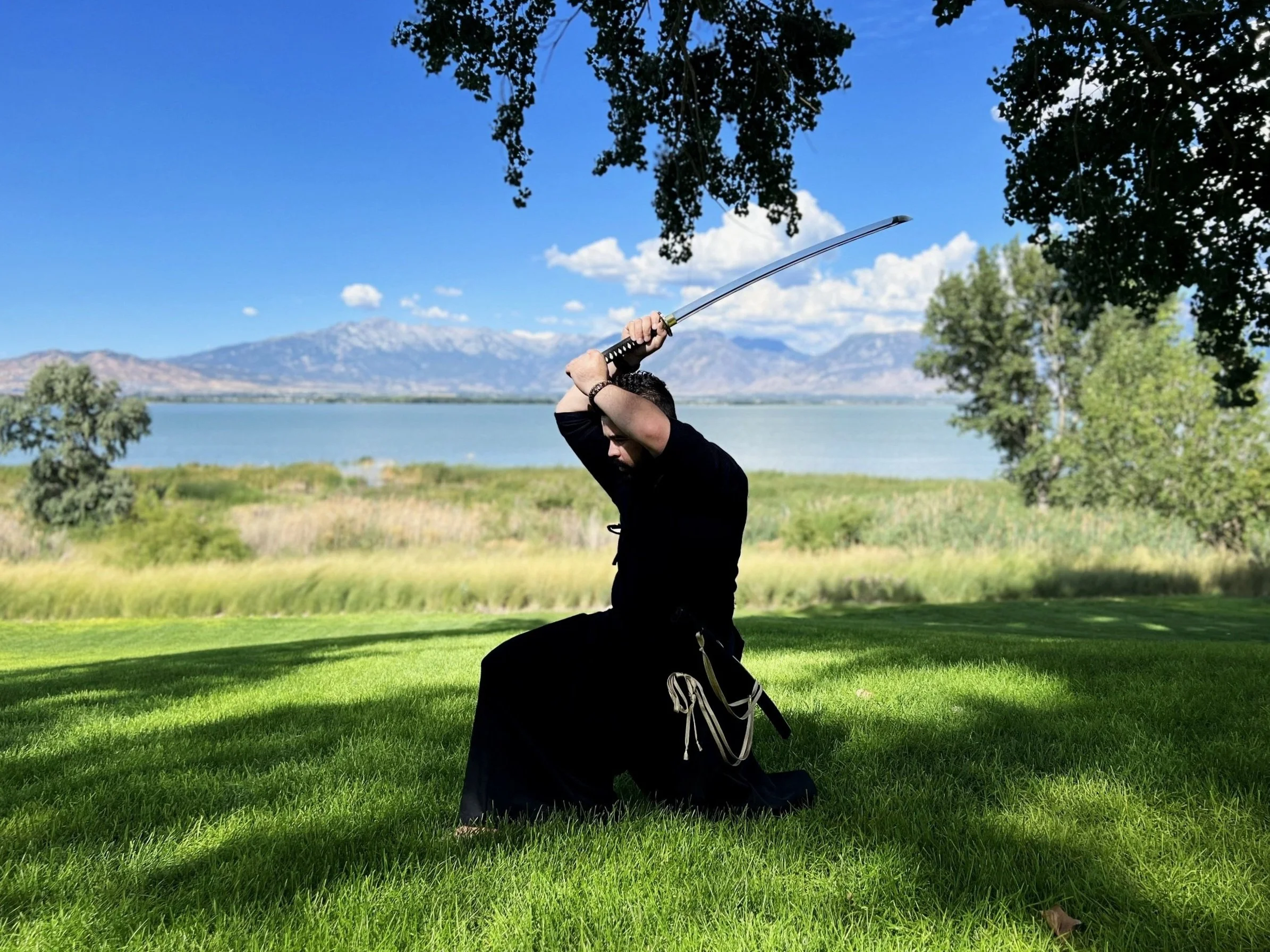 Person kneeling on grass, holding a katana sword, outdoors with mountains, lake, trees, and blue sky in the background.