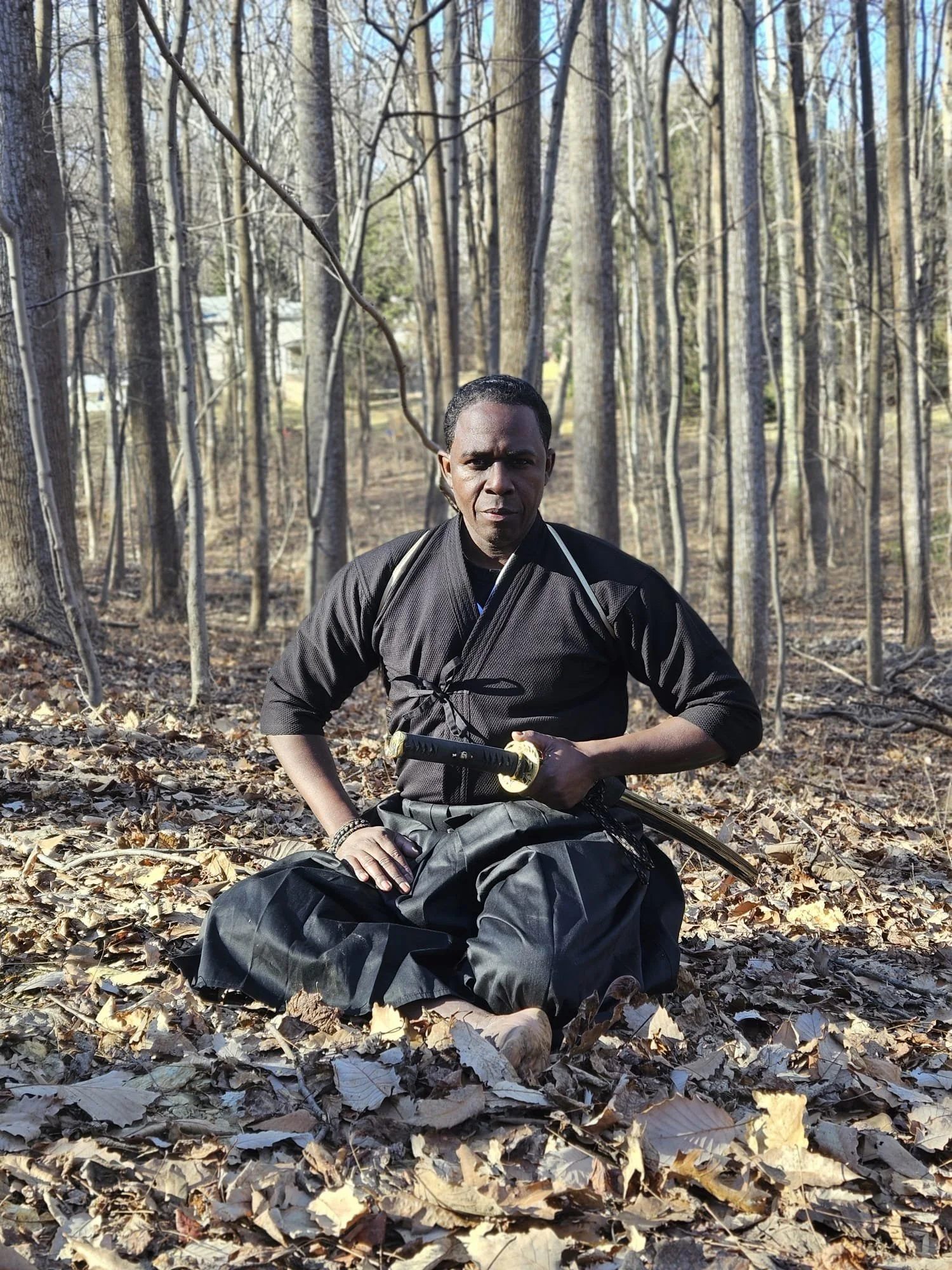 A man dressed in martial arts attire, sitting on the ground in a forest with fallen leaves, holding a sword.
