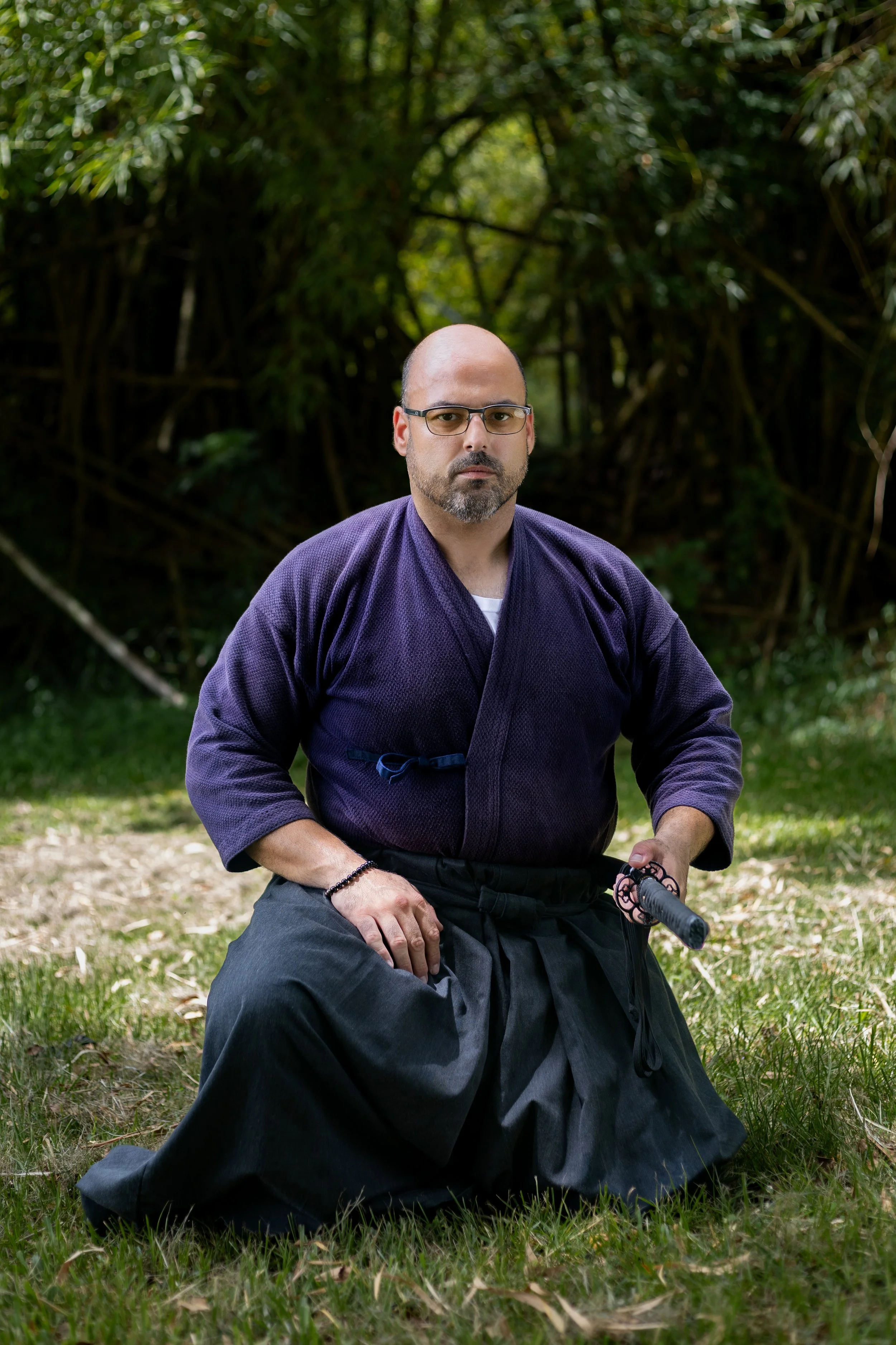 A man kneeling on grass, dressed in a purple martial arts gi and black hakama, holding a small sword or dagger, outdoors with dense green foliage and a wooden sign with Japanese characters in the background.