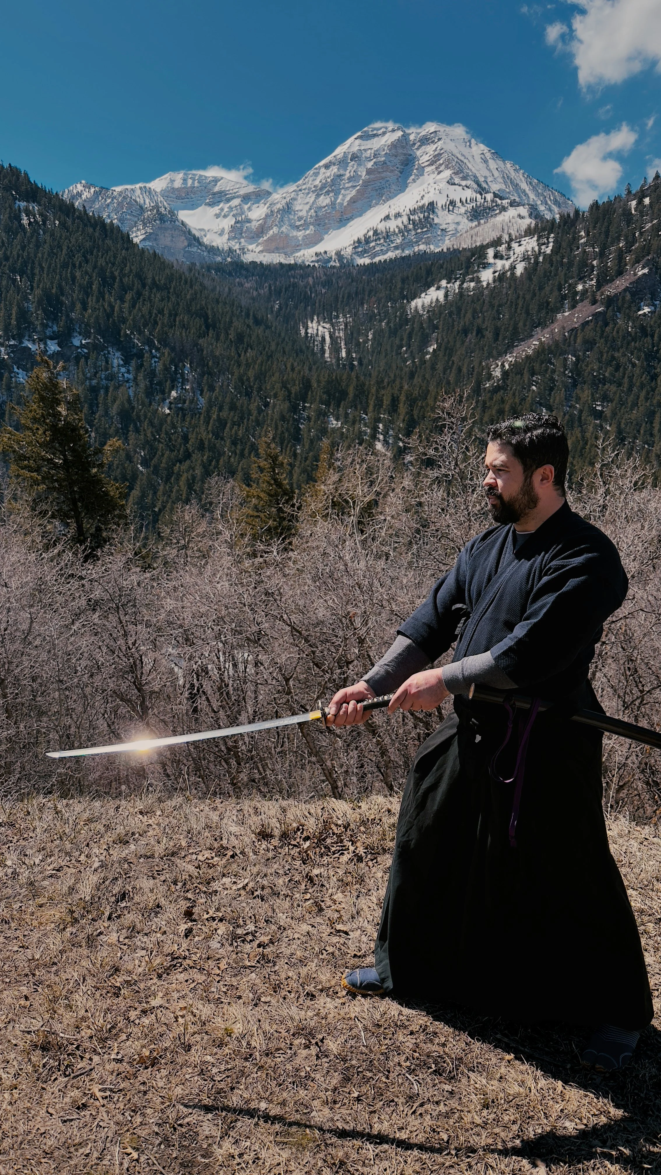 A man in traditional martial arts clothing practicing with a sword outdoors with mountains and trees in the background.