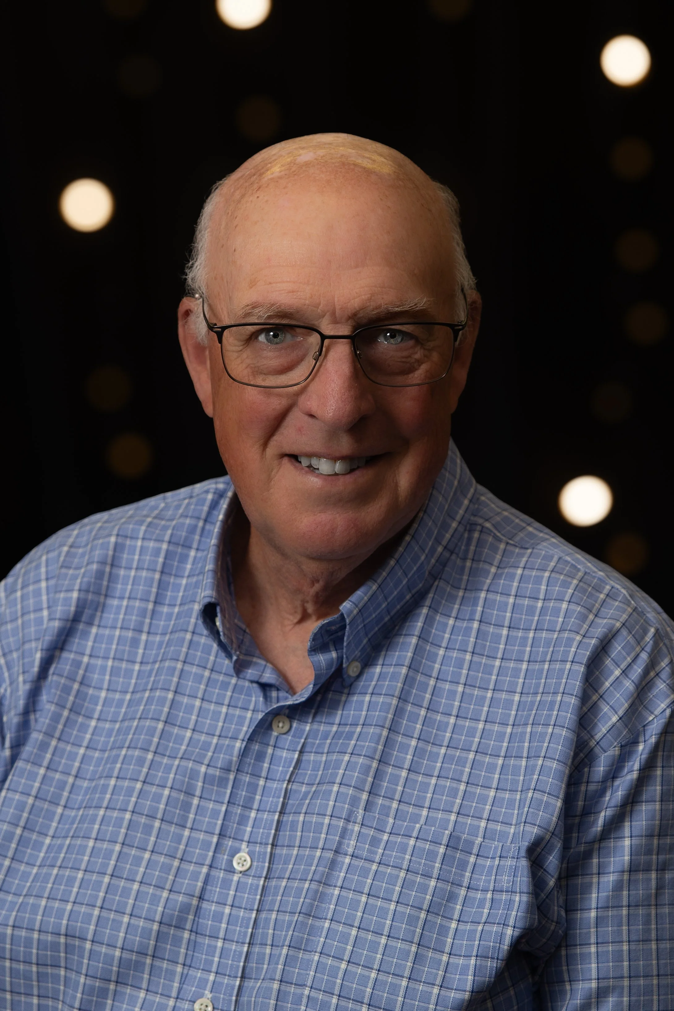 A smiling elderly man with glasses, wearing a checkered shirt, standing in front of a brick wall.