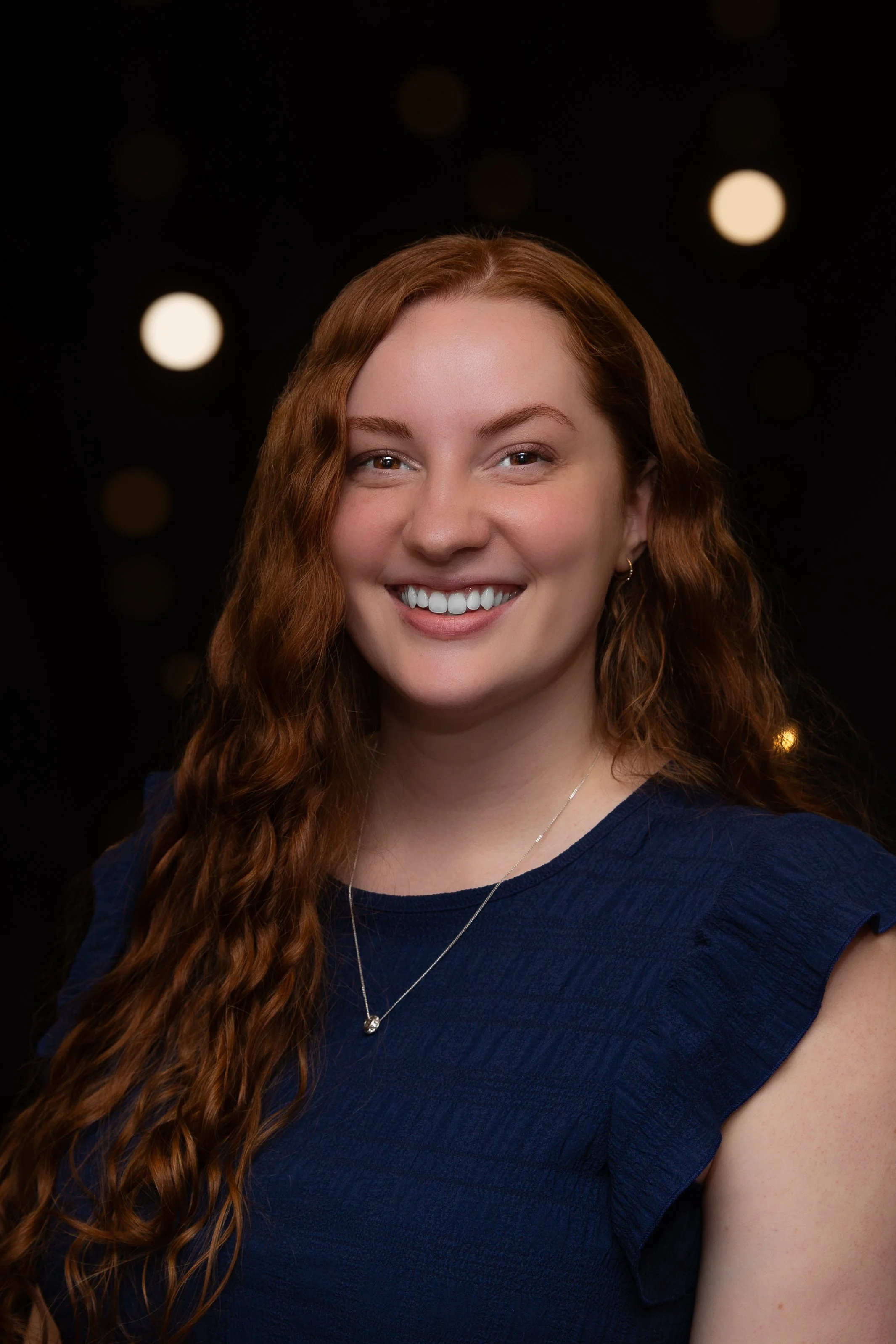 A young woman with red wavy hair and fair skin smiling outdoors, standing in front of a wooden fence with green foliage and sunlight shadows.