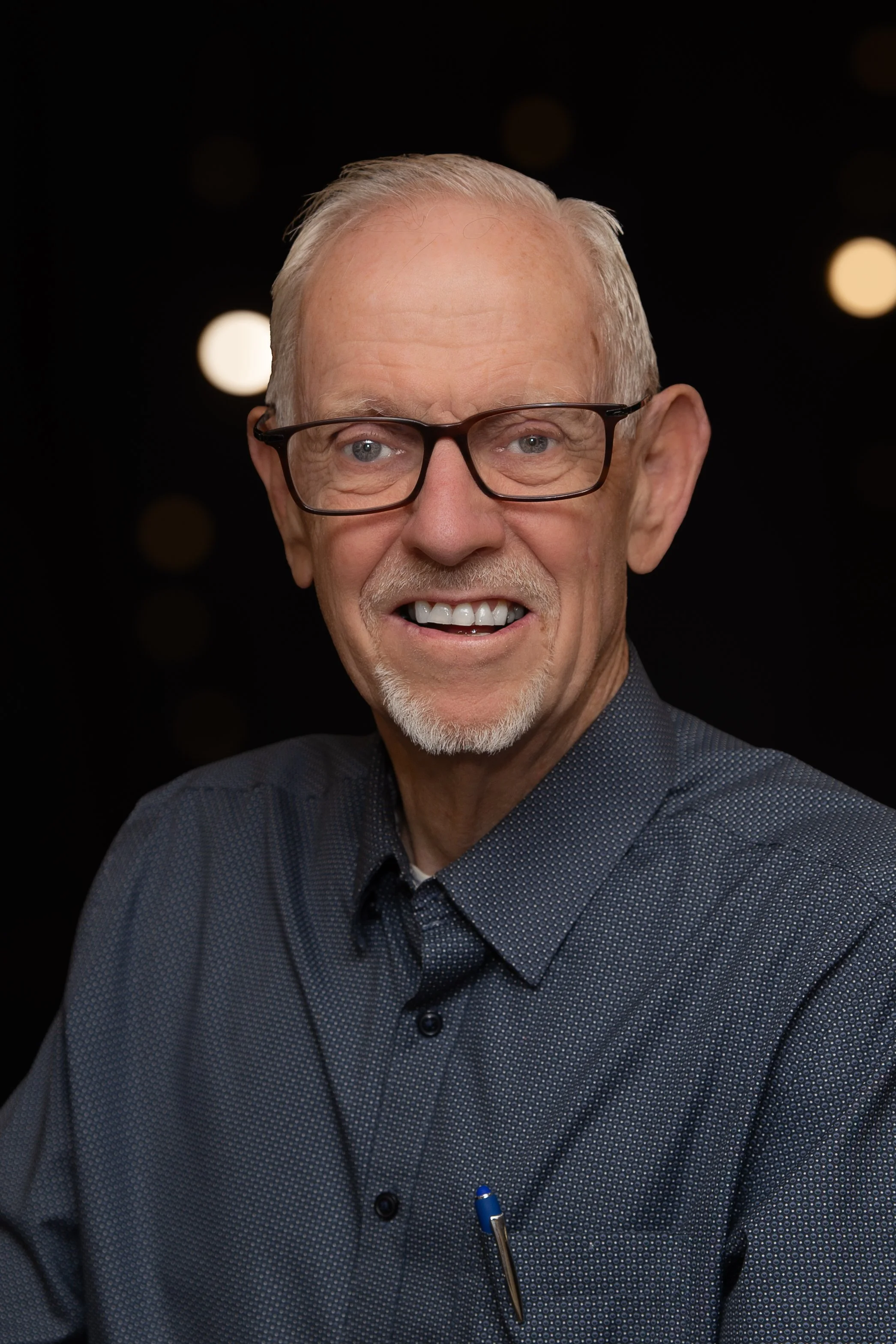 Smiling elderly man with white hair, glasses, and a beard, wearing a plaid shirt, in front of a brick wall.