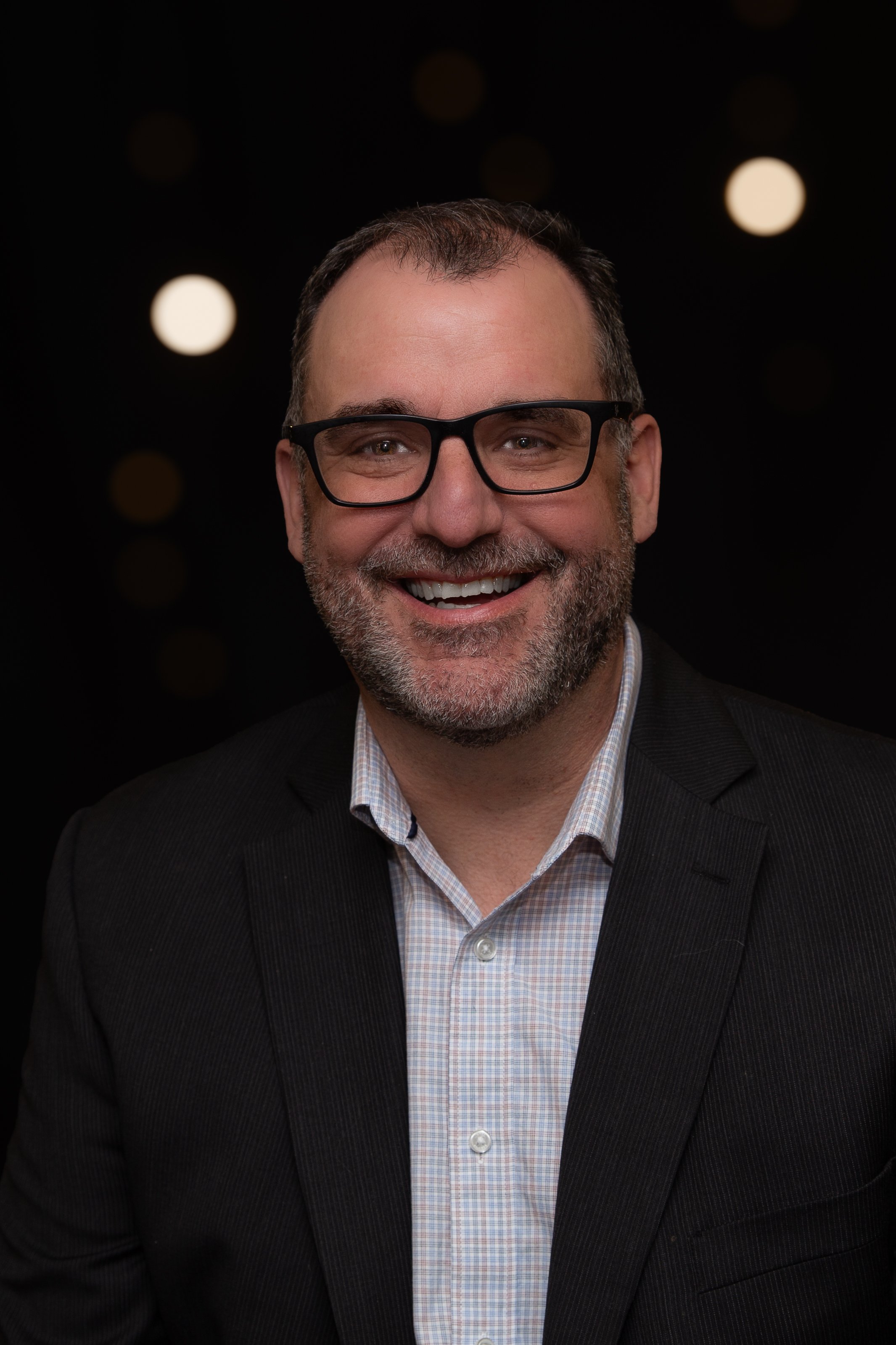 A smiling man with short dark hair and a beard, wearing a dark blazer and blue shirt, standing in front of a wooden wall with warm lighting.