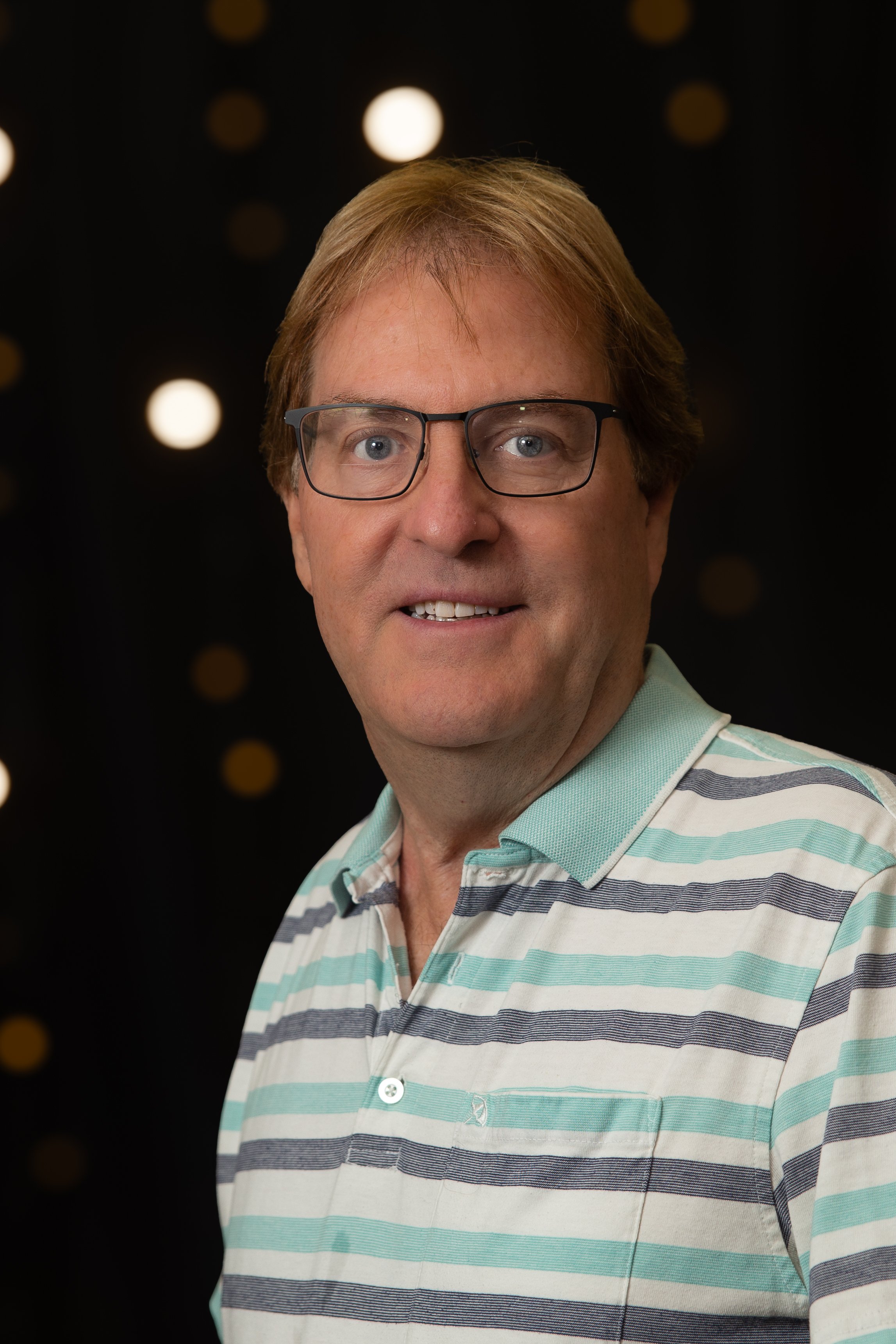 Close-up of a man with glasses, light brown hair, and a friendly smile, indoors with blurred background.