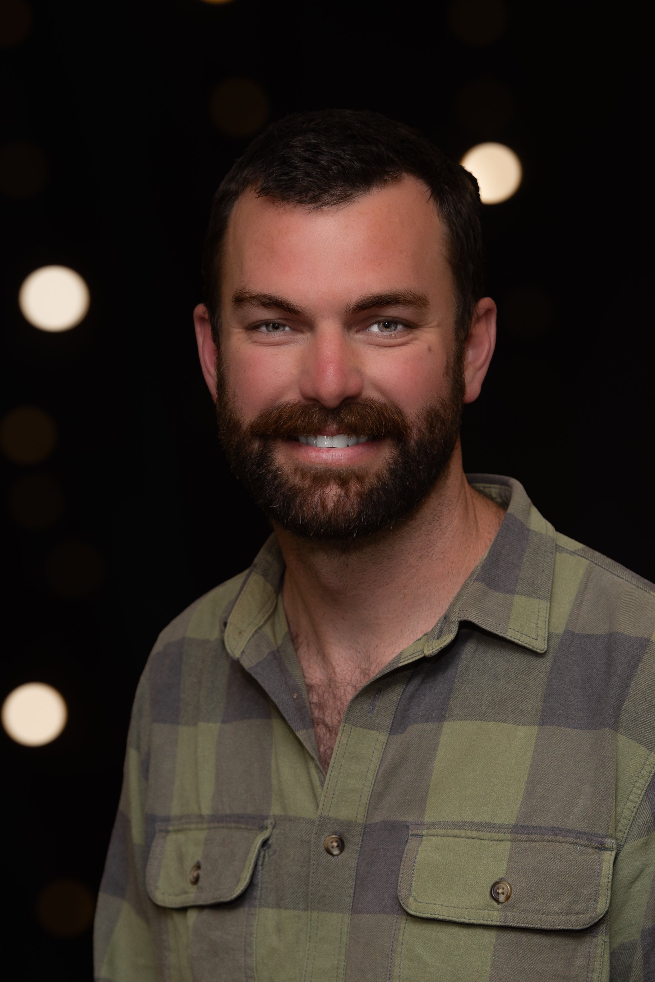 Close-up of a smiling man with brown hair, a full beard, light skin, and blue eyes, standing in front of a wooden background.