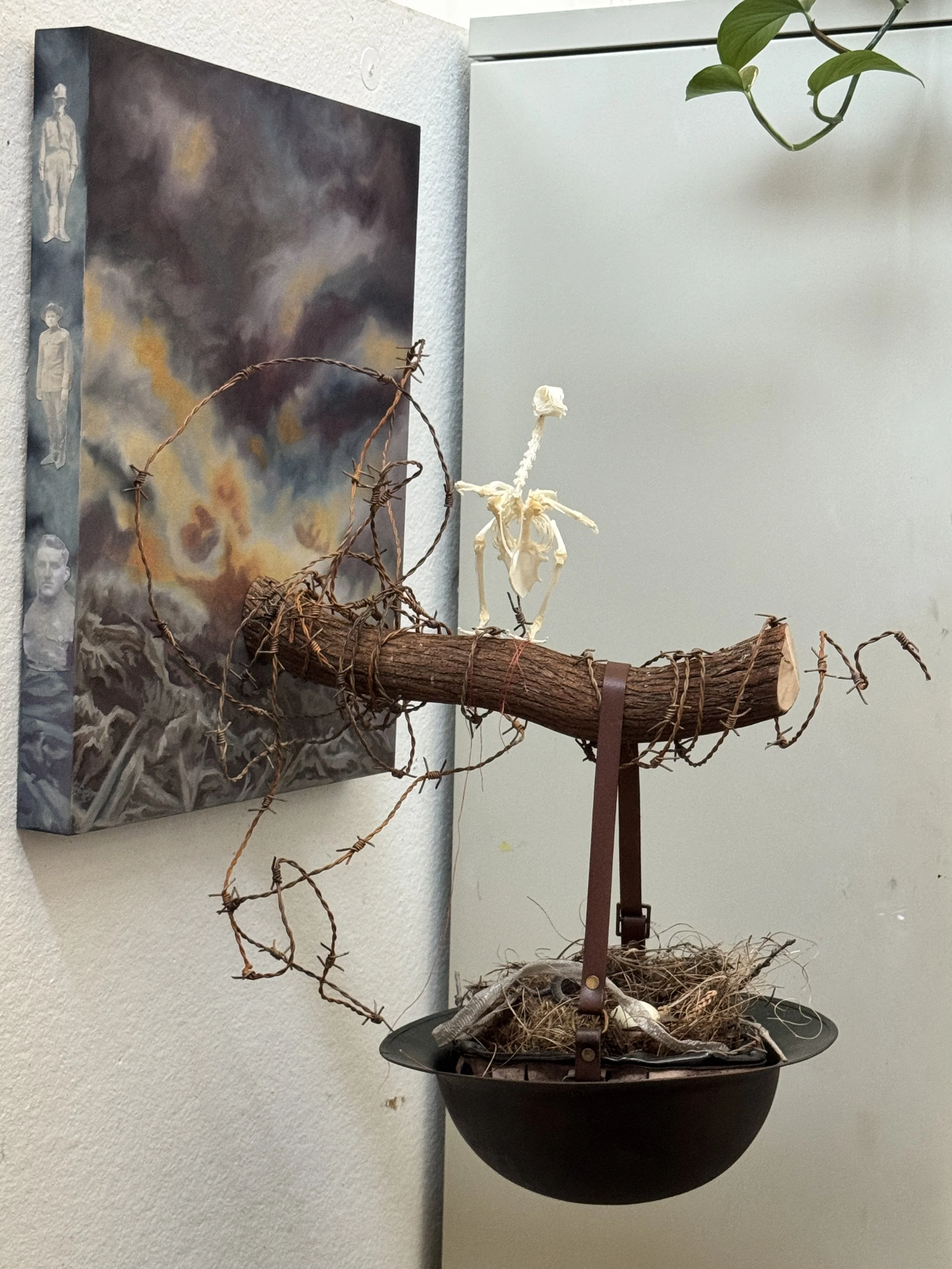 Ghosts of World War One. Mixed Media Assemblage: Oil on wood, photos, branch, rusted barbed wire, red thread, pigeon skeleton, replica WW1 helmet, found nest, replica pigeon egg, mummified snake, shed snake skin. 2025.