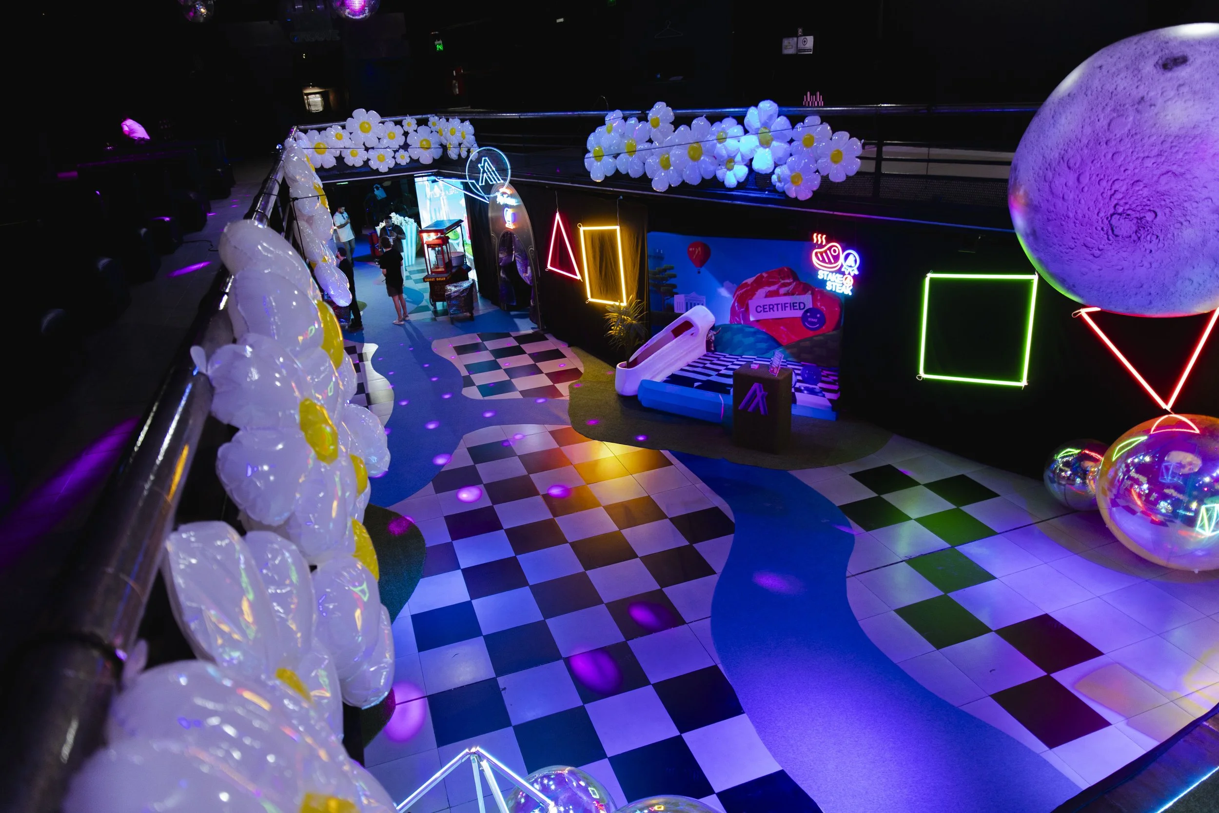 A colorful indoor space with neon lights, balloon decorations, a bed with pillows, and various playful decor elements, resembling a modern, vibrant play or lounge area.