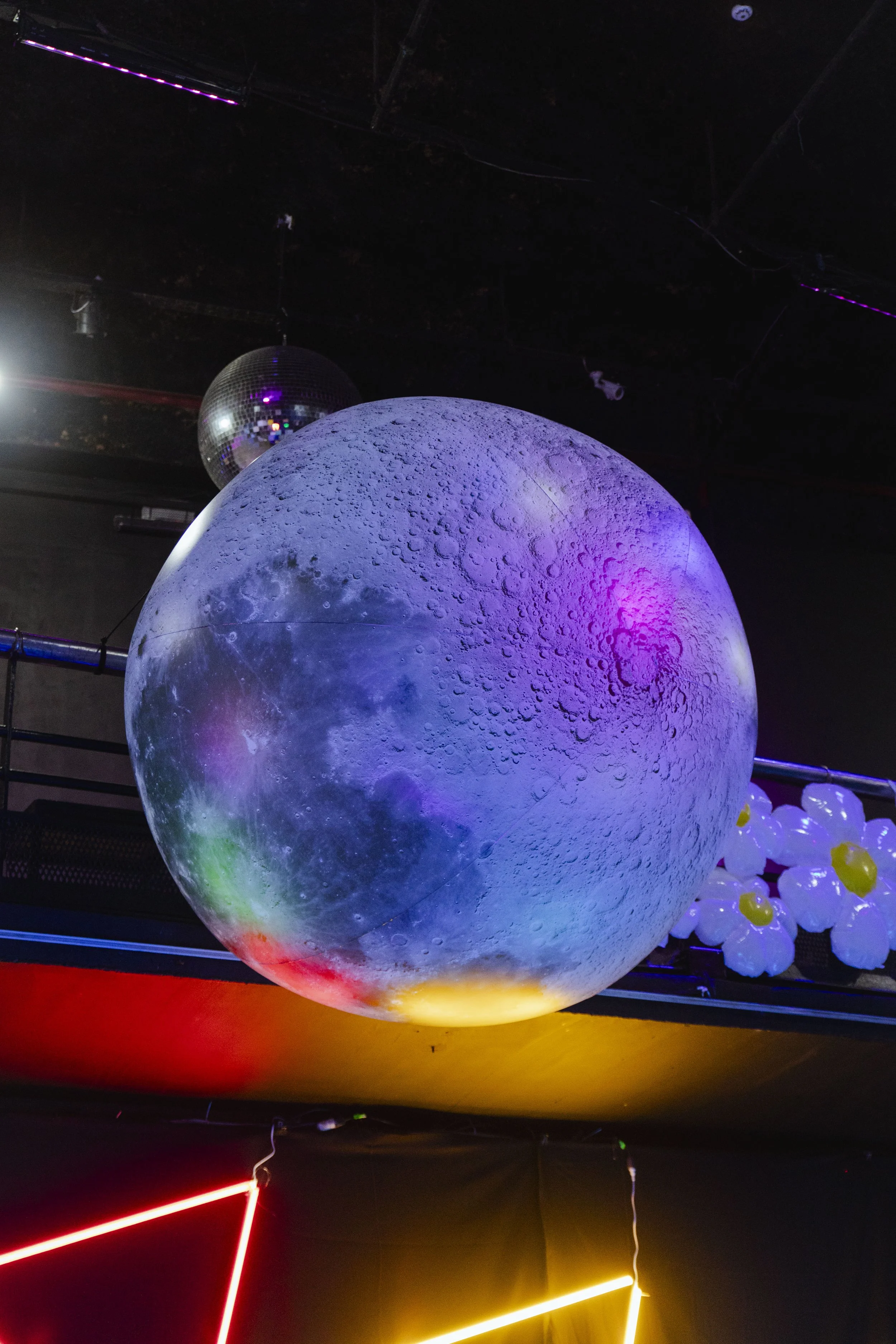 A large moon and disco ball hanging from a ceiling, with colorful lighting and balloon decorations nearby.