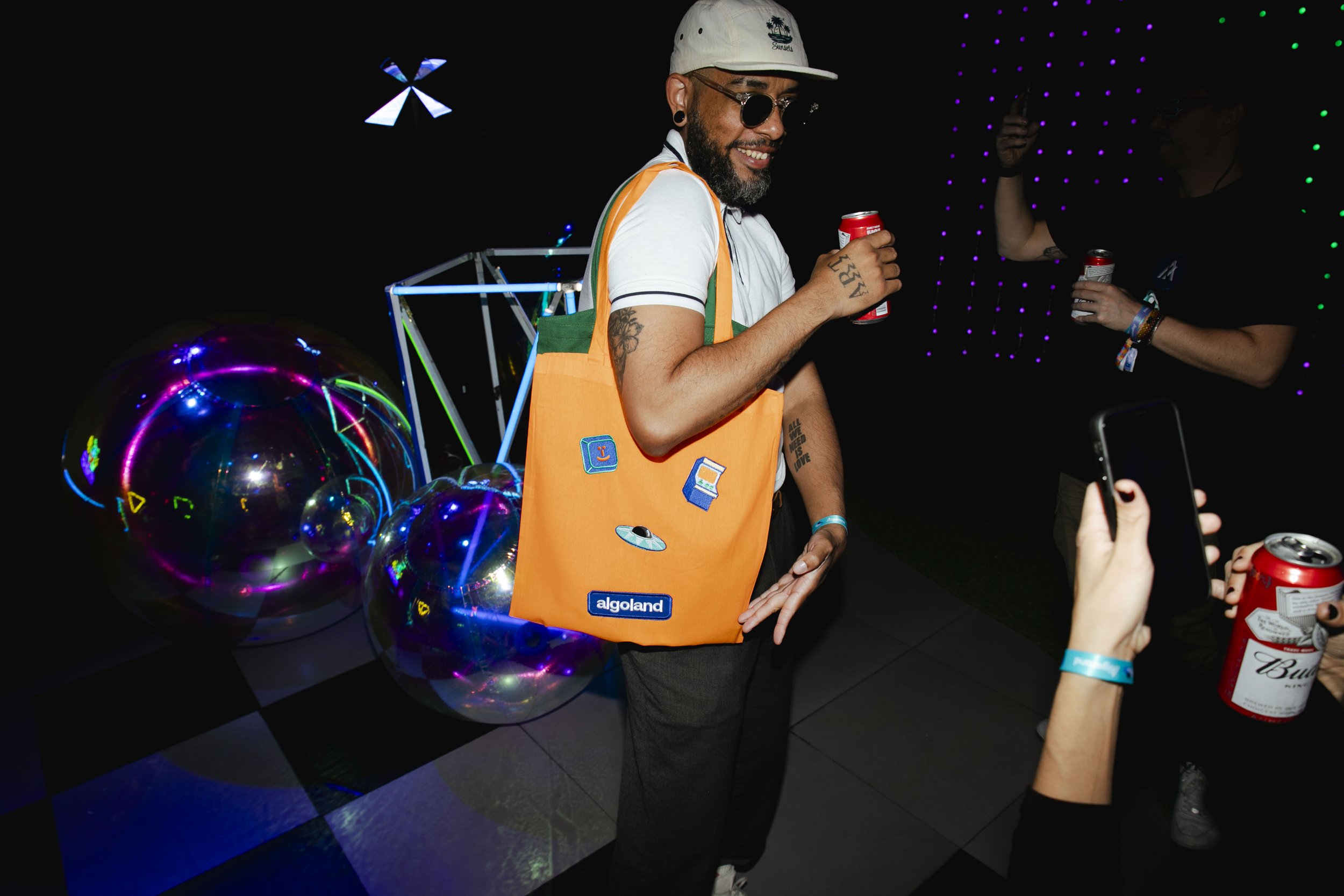 Man with tattoos, sunglasses, and a white cap smiling at a party, holding a red can, wearing an orange tote bag with patches, and standing in front of reflective balloons with neon lights and other people holding cans in a dark setting.