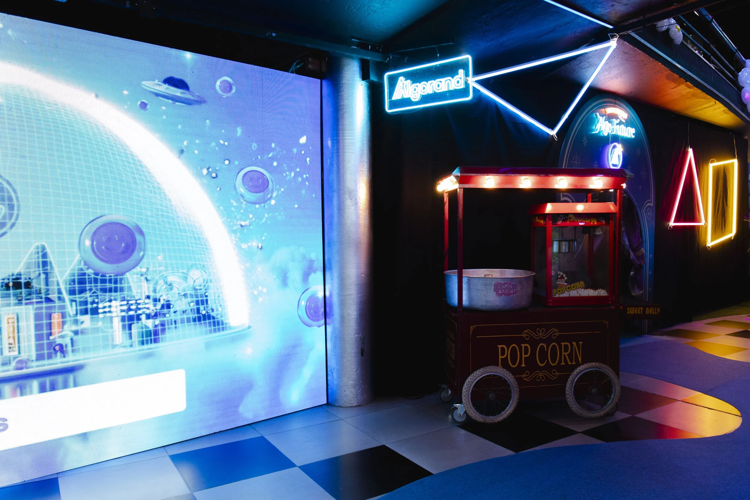Indoor scene with neon signs, a popcorn cart, and a large digital screen displaying a futuristic graphic with planets and a grid, with checkered flooring and decorative lighting.