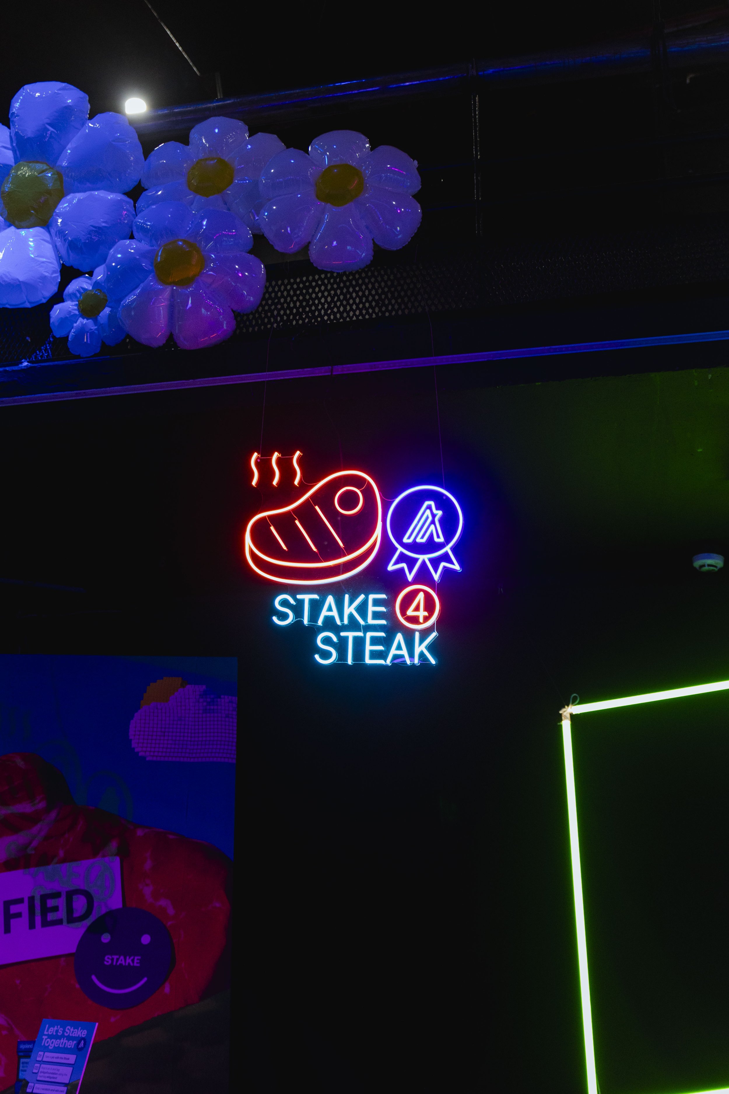 Neon sign that reads 'STAK 4 STEAK' with a steak graphic and award ribbon, hanging below a cluster of illuminated white and yellow flower decorations.