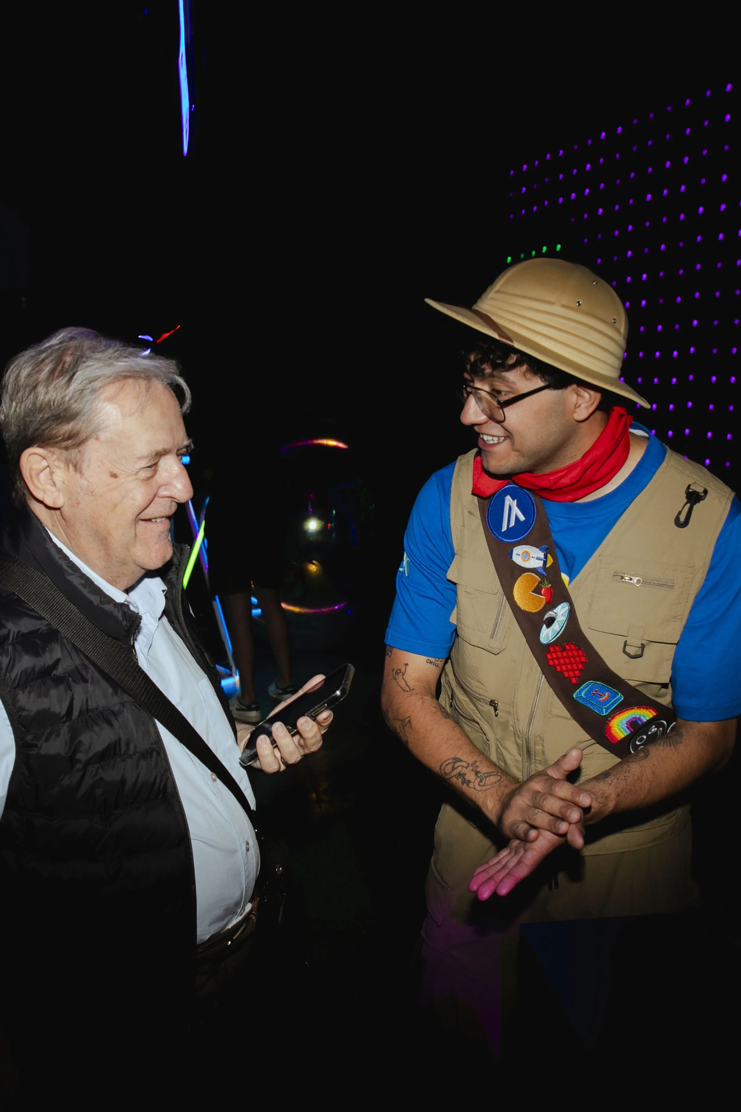 A man in a blue shirt and camera badge talks to a man in a black vest holding a smartphone, both smiling, in a dark room with colorful neon lights.