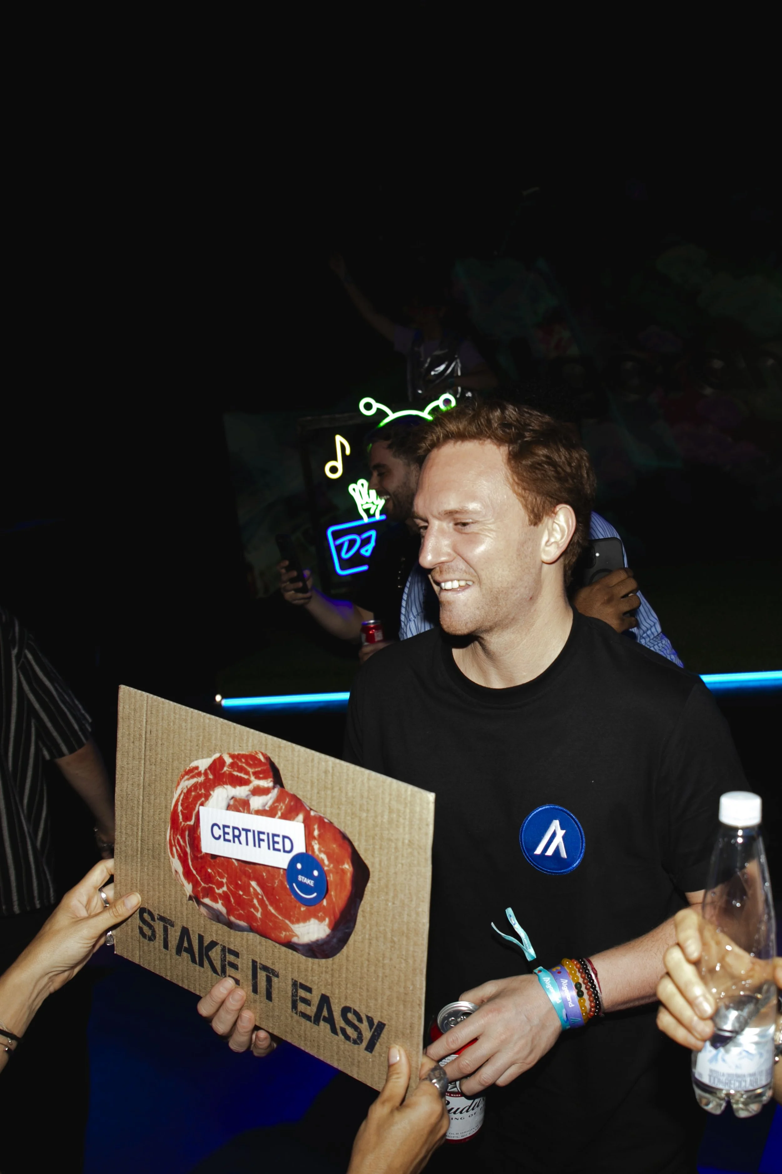 A man with red hair smiles while holding a cardboard sign that says 'STAKE IT EASY' with a picture of a cut of steak saying 'CERTIFIED'. A hand holds a bottle of beer, and another hand holds a water bottle. There are festive neon lights in the backgr
