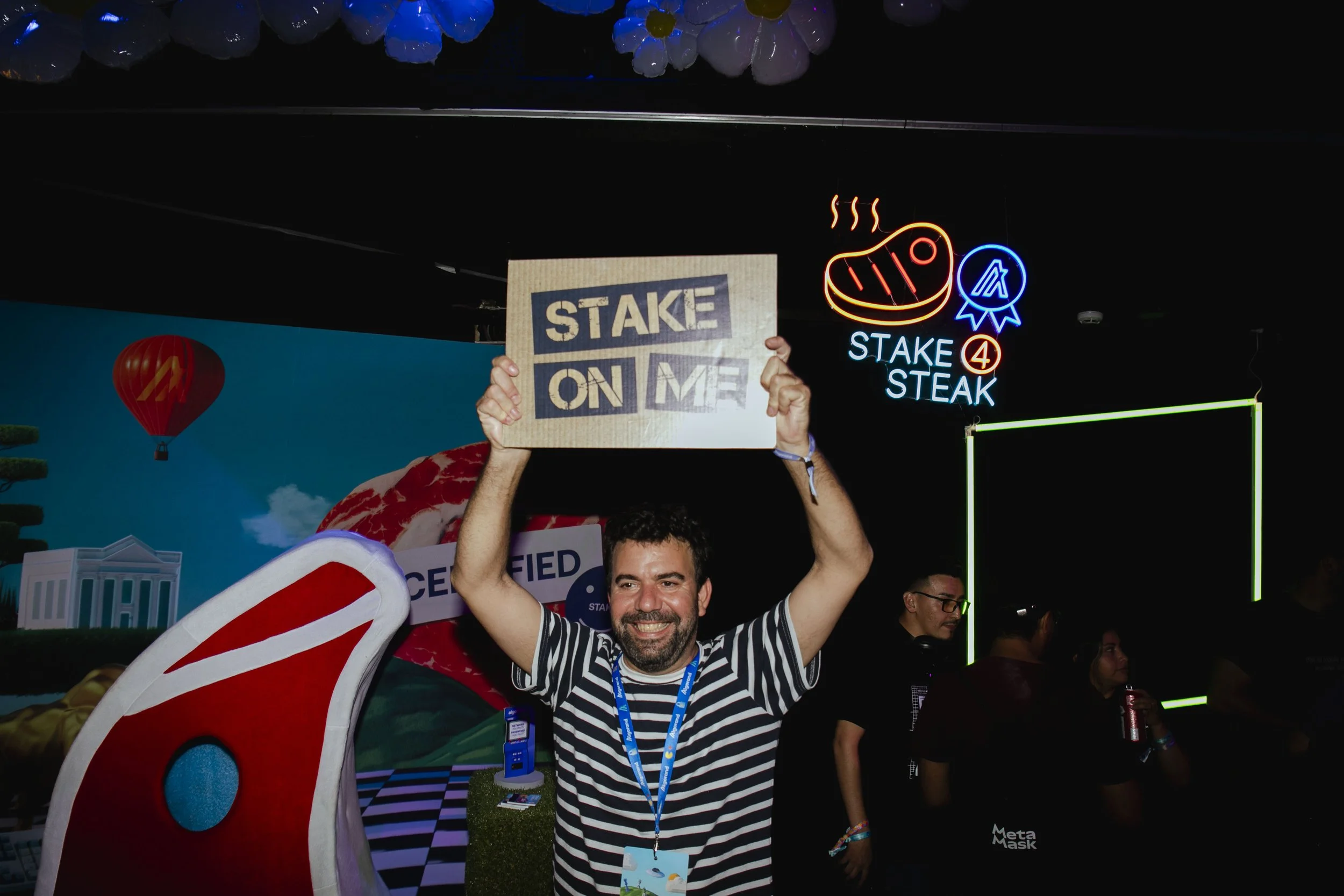 A man in a striped shirt smiling while holding a cardboard sign that says 'STAKE ON ME' at an event with neon signs, including a steak icon, in the background.